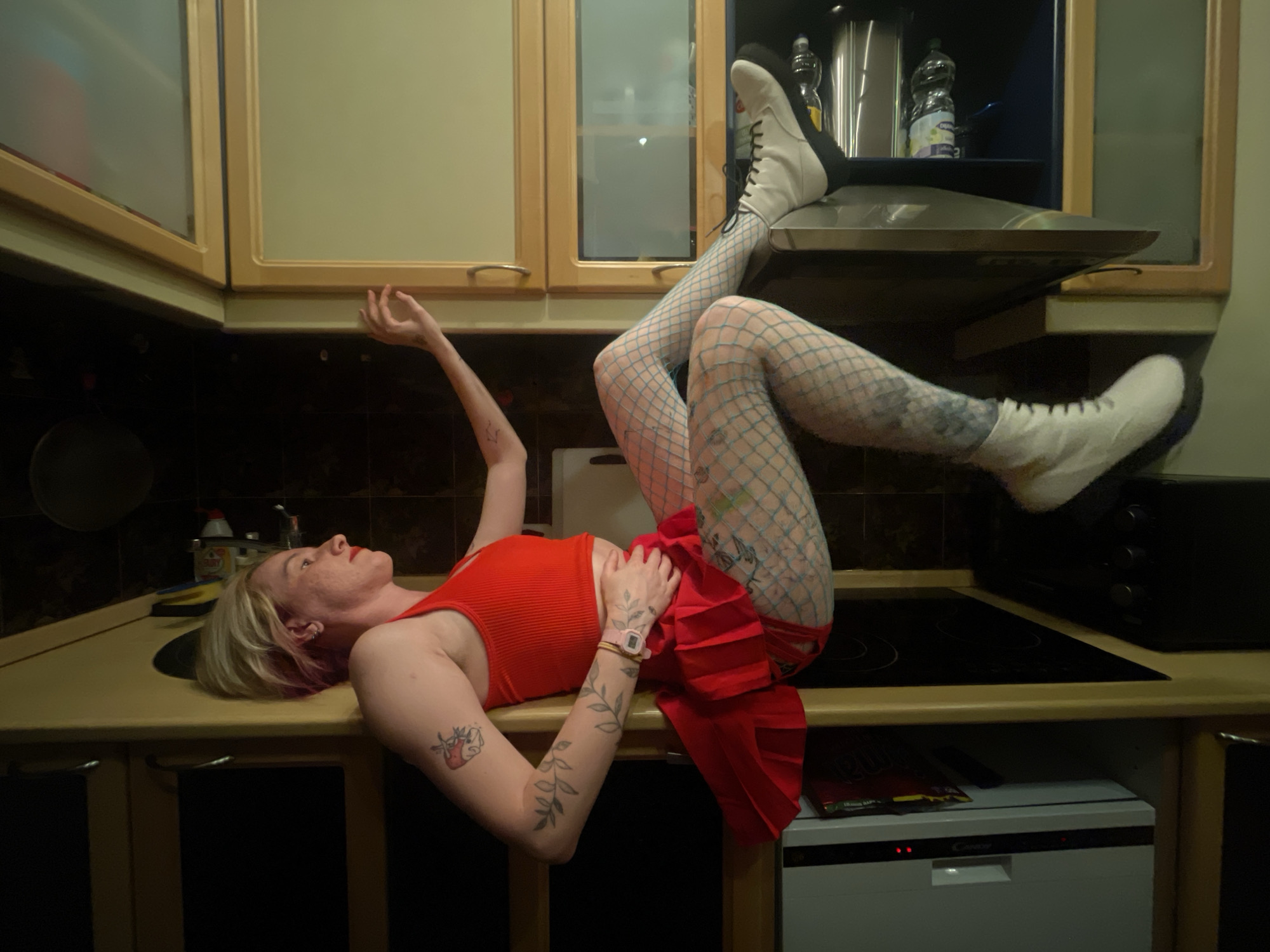A woman lies on a kitchen counter. She is wearing white boots with black soles, blue fishnet stockings, a red dress, and a red top. She has tattoos on her arms and legs. The kitchen has cabinets, a stovetop, and a dishwasher. The location is likely within an apartment in the city of Vracar Urban Municipality, Serbia.