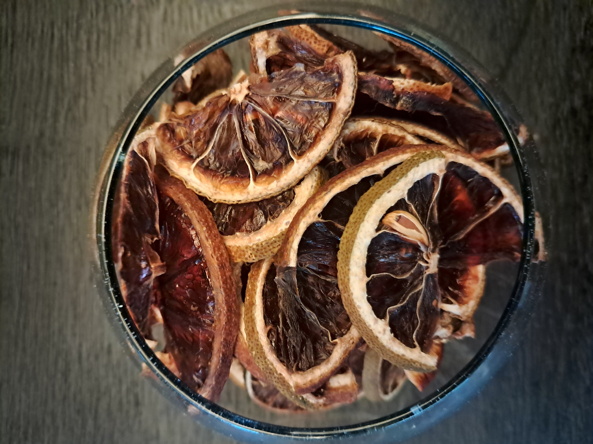 A top-down, close-up shot shows a clear glass bowl filled with dried citrus slices. The slices, likely oranges or grapefruits, are arranged haphazardly, filling the bowl. The fruit's flesh appears dark brown and shriveled, while the rinds are a lighter tan with some green. The bowl is sitting on a dark gray surface. The exact location within Vicente López, Argentina, cannot be determined from the image.