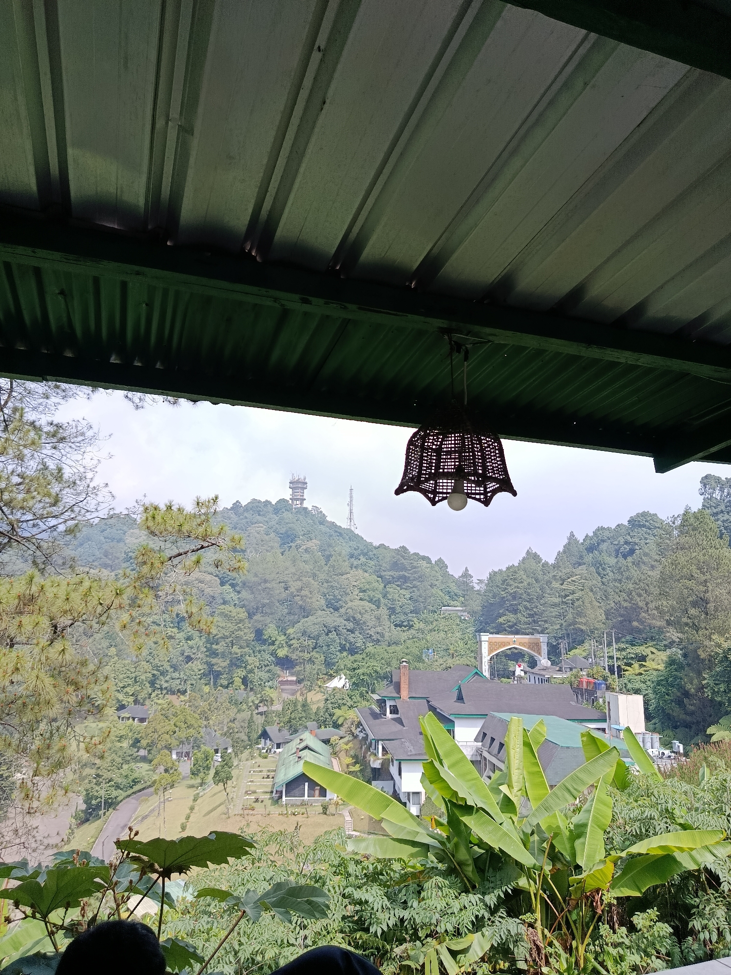 The image depicts a view from a covered outdoor seating area, looking out over a lush, hilly landscape. The foreground features large green leaves, possibly from banana plants, and the dark silhouettes of at least two people sitting in the lower portion of the frame. Above, a corrugated metal roof with a green underside is visible, from which a woven basket-style lampshade hangs.

The midground shows a sprawling resort or hotel complex nestled into the hillside, with buildings featuring dark roofs and white walls. An arched gateway is visible in the distance. The background is dominated by dense, green forest covering rolling hills. A prominent tower, likely a telecommunications tower, stands on the highest peak in the distance, with smaller antennae visible nearby. The sky is overcast, suggesting a slightly hazy or cool day. The overall impression is of a tranquil, scenic location, possibly a mountain resort area. The scene is consistent with locations found in the mountainous regions surrounding Cianjur, Indonesia, known for their natural beauty and tourist destinations.