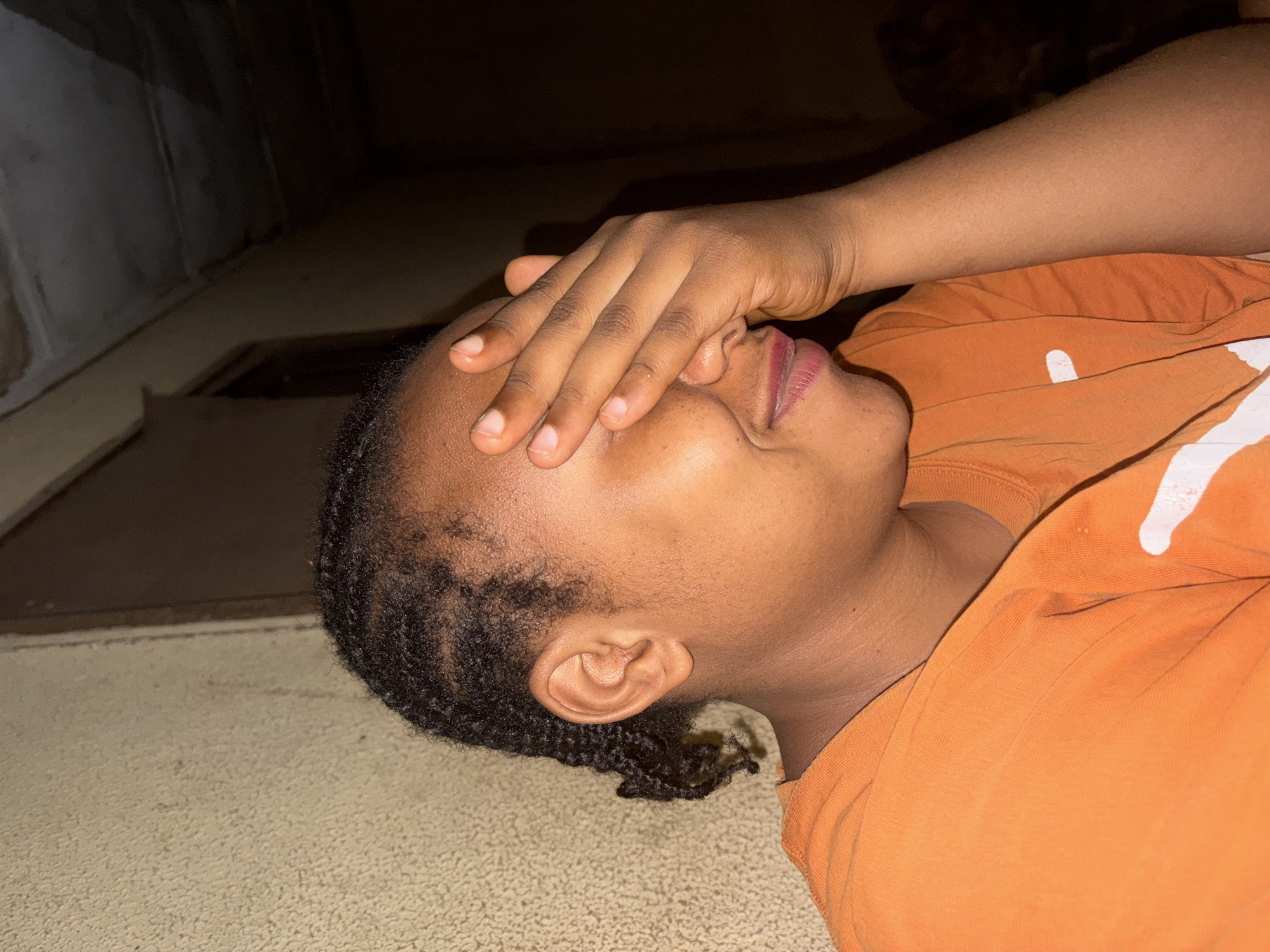 A person lies on their side in a dimly lit indoor setting in Jos, Nigeria. The individual has dark skin, with their head resting on a light-colored, textured surface, possibly a floor or mat. Their hair is dark and styled in short braids or natural curls. They are wearing a bright orange t-shirt, which has a prominent white, abstract design or mark visible on the chest area.
The person's right hand is placed over their face, covering their eyes and forehead, with fingers spread slightly. Their lips are slightly parted, revealing a hint of a smile or a relaxed expression beneath the hand. Only a portion of their face, from the nose down to the chin, and their right ear are clearly visible. The background is dark and indistinct, suggesting a corner of a room or a wall in low light. The overall atmosphere is quiet and subdued, with the low lighting creating a sense of intimacy or rest. The individual's posture suggests a moment of relaxation, perhaps shielding eyes from a light source, or a gesture of contemplation or shyness.