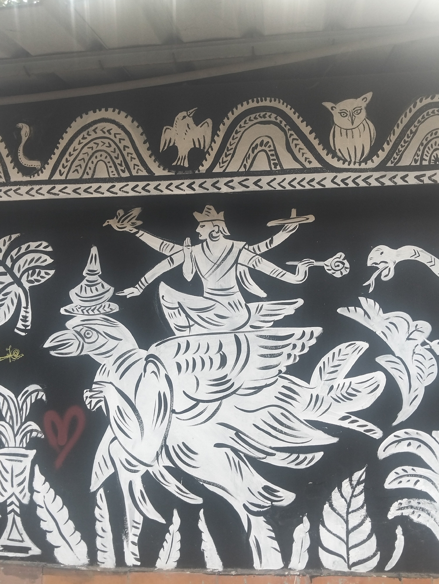 The image is a black and white mural on a wall, possibly in Dhaka, Bangladesh, depicting a mythological scene. In the upper register, there is a decorative border featuring a snake, a bird resembling an eagle, and an owl. Below this, a four-armed figure sits atop a large bird with substantial plumage. The figure holds a bird in one hand, and possibly a flower or other object in another. Opposite the bird is a cobra. Additional stylized plants and trees are depicted. A red heart is sprayed on the wall in the lower left. The mural is near the ground, possibly at the base of a wall.