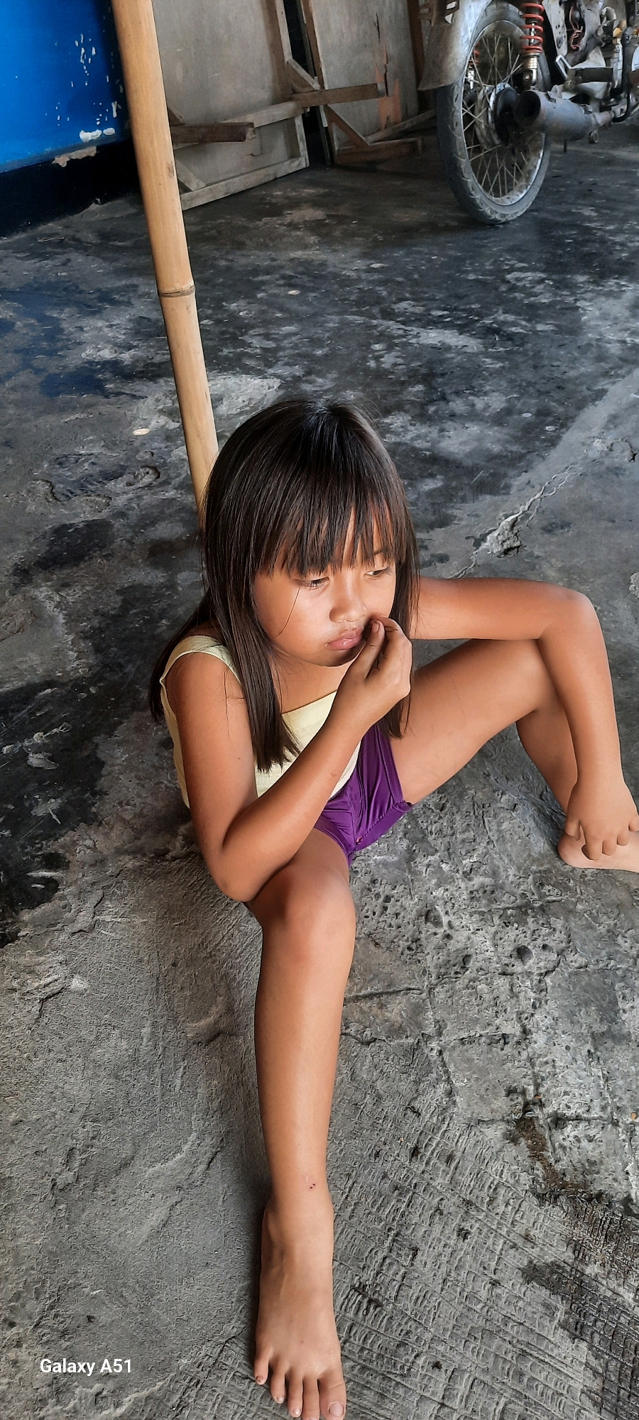 A young girl with dark hair is sitting on a concrete floor. She is wearing a light yellow sleeveless top and purple shorts. She is looking down and has her finger to her mouth, appearing to be deep in thought or perhaps a little bored. A bamboo pole is leaning nearby, and behind her, there are some wooden structures and a motorcycle. The ground is rough and textured, typical of an outdoor or semi-outdoor setting. The overall impression is one of a candid moment, possibly in a village or rural area in Indonesia, given the location context. The lighting suggests it could be daytime. The bottom left corner of the image shows the text "Galaxy A51," indicating the device used to capture the photo.