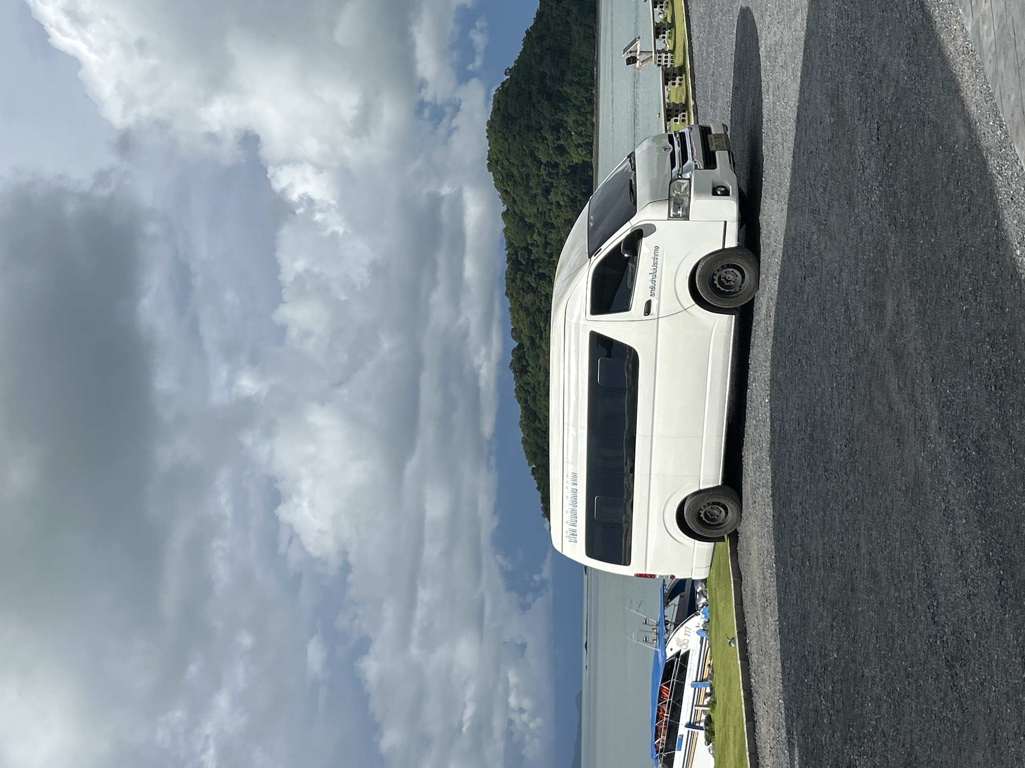 A white van is parked on a gravel lot next to a body of water. The van has tinted windows and text written on its side in Thai. Several boats are docked in the water behind the van. In the distance, a lush, green island rises from the water. The sky is filled with dramatic, gray clouds. The scene appears to be in Ratsada, Thailand, a coastal area known for its beautiful scenery and boat tours.