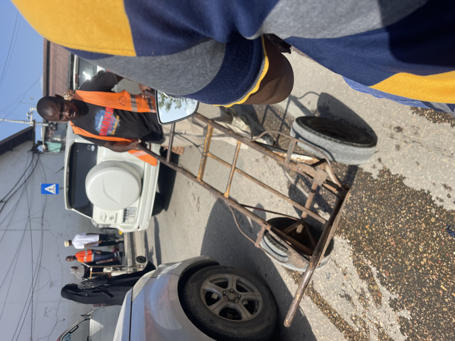 An urban street scene in Zanzibar, Tanzania, is depicted under direct sunlight. In the midground, a Black male wearing a black t-shirt, an orange safety vest, and light-colored pants is pulling a rusty, four-wheeled hand cart forward. The cart is empty. To the immediate left of the cart is the front wheel and lower portion of a silver or light-colored vehicle. Behind the man with the cart, a white Toyota Rav4 SUV is parked, identifiable by its rear-mounted spare tire and a partially visible Tanzanian license plate "T 933 LFG". In the background, on the left side of the street, several individuals are visible: one person in a black abaya, another in an orange vest, and a third in a white shirt and head covering, among others. Buildings line the street, and overhead power or communication cables are present. A blue road sign with a white pedestrian icon is mounted on a pole above the white SUV. The foreground on the right shows the shoulder and arm of an individual wearing a blue and yellow striped shirt, with a motorcycle mirror reflecting the sky and trees. The asphalt road surface exhibits dark, wet-looking patches.