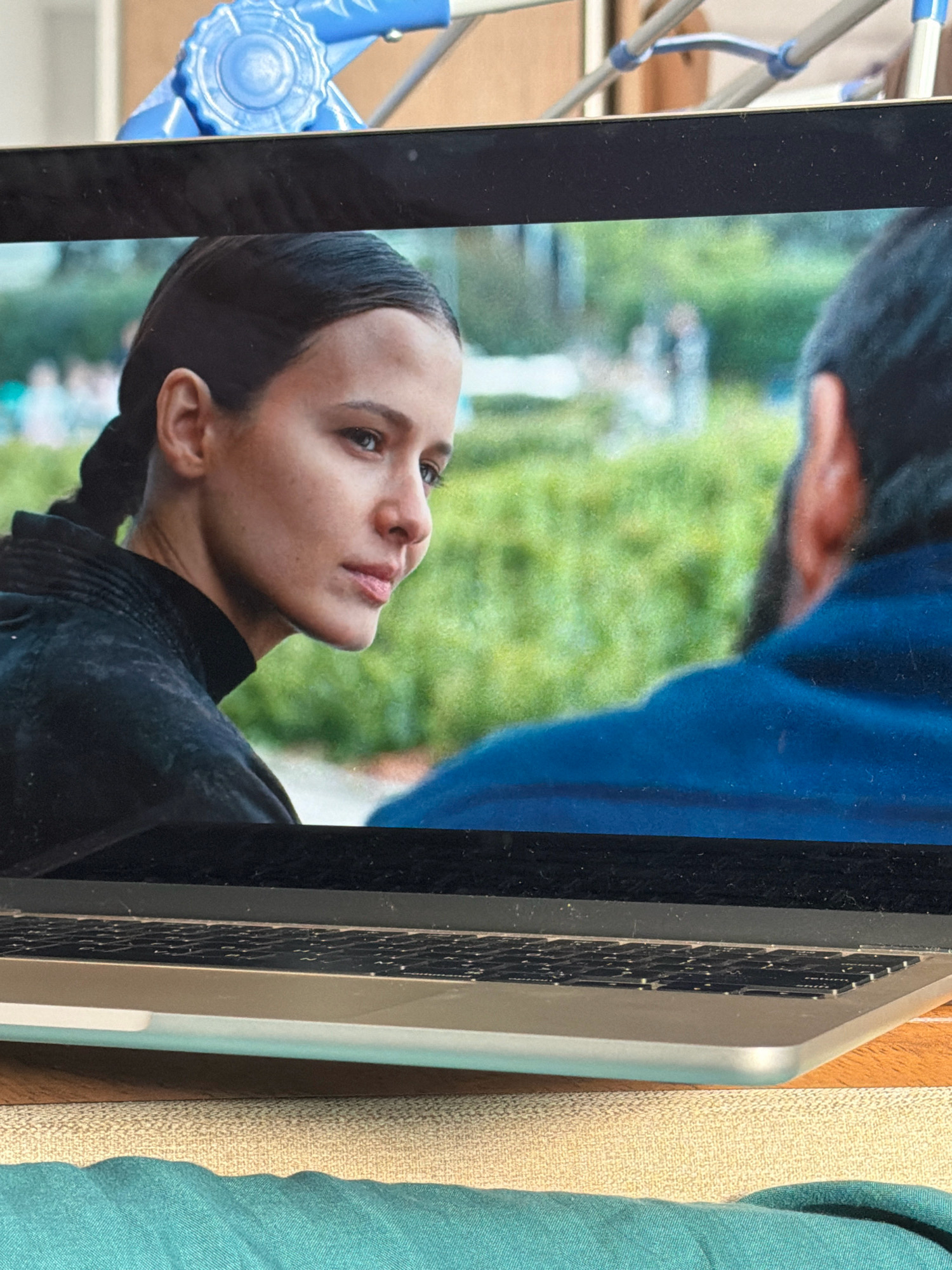 The image shows a laptop screen displaying a close-up of a woman with dark hair pulled back in a braid, wearing a black top. She is looking to the right with a serious expression. In the background of the screen, out of focus, is a man in a blue shirt and a blurred outdoor setting with greenery. The laptop is positioned on a table, with the keyboard visible in the foreground. Above the laptop, a blue object with circular detailing is visible, possibly part of a rack or stand. The scene is indoors, with the laptop resting on a textured surface, possibly a table or desk, and a turquoise fabric is draped in the lower part of the image. There are no explicit indicators of the location being Khabarovsk, Russia.