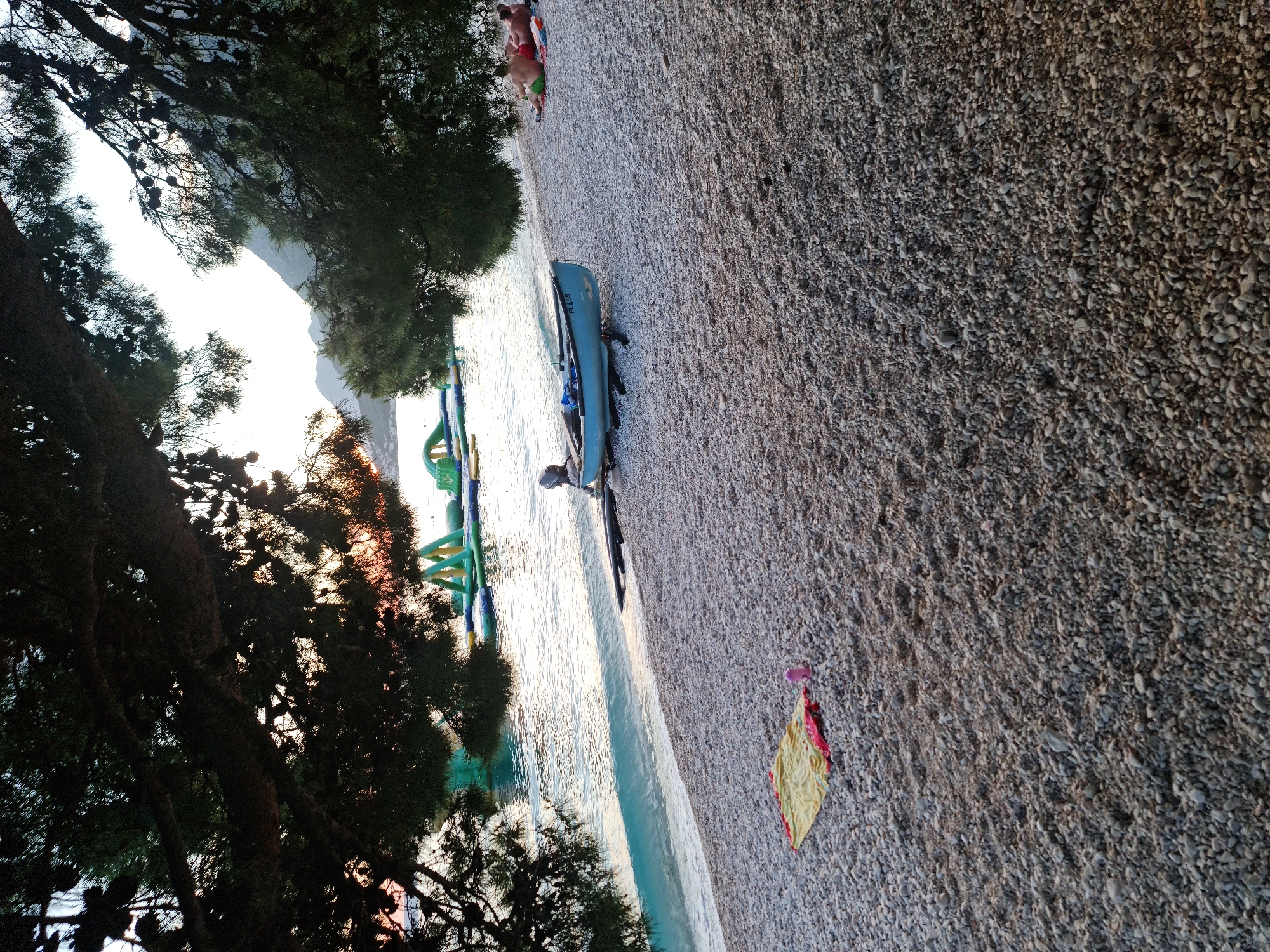 A pebble beach in Općina Gradac, Croatia, features a blue boat resting on a trailer near the water's edge. The water is calm and a light turquoise near the shore, with a floating inflatable water park visible in the distance. A yellow and red towel with a pink object lies on the pebbles in the foreground. Two people are relaxing on towels on the beach to the right. Overhanging tree branches with pine cones frame the top of the image, casting shadows over the scene and obscuring the setting sun, which creates a warm glow on the horizon.