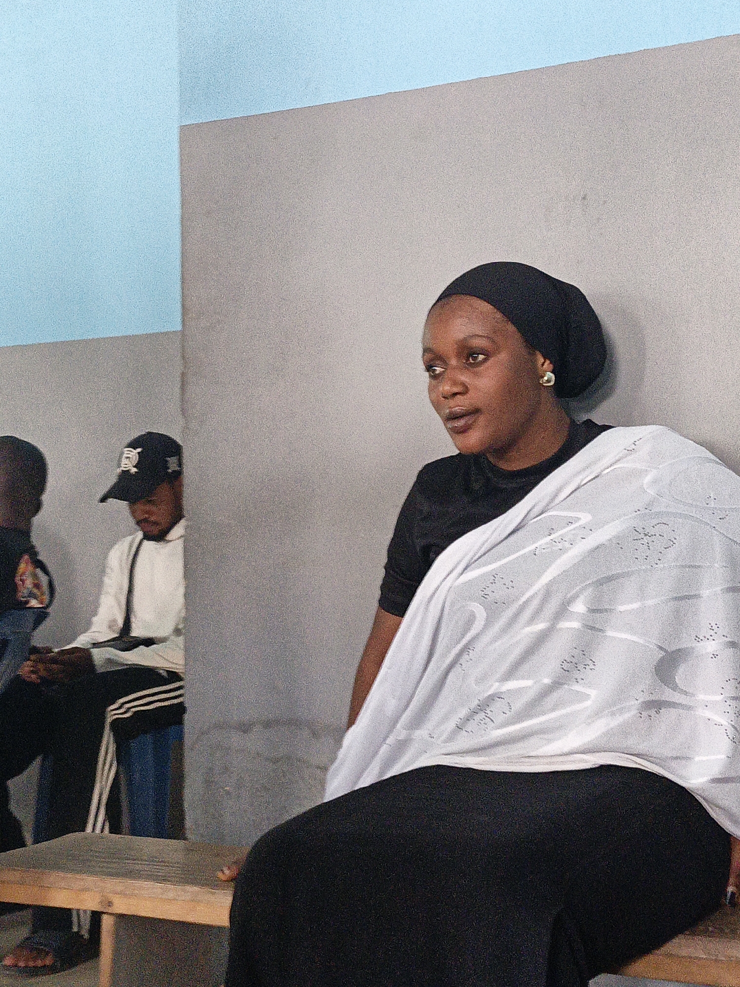 A medium shot shows a woman seated on a wooden bench in what appears to be an interior room. The woman has dark skin and is wearing a black head covering, a black top, and a long black skirt or dress. A white shawl or garment is draped over her shoulders. She is looking to her left, away from the camera.
In the background, partially obscured, are two other individuals. One is wearing a black baseball cap and a white jacket, and the other is wearing a dark garment. They are also seated. The room has walls painted in a light blue and gray color scheme. The location is in the city of Fagge C, Nigeria.