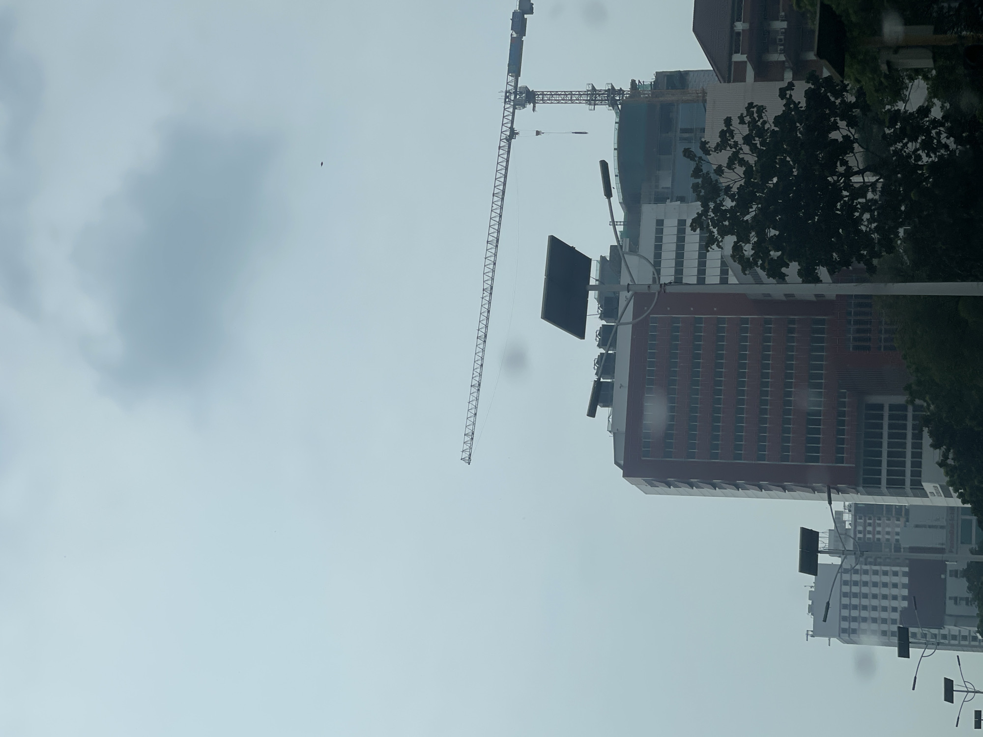 The image depicts a view of a city skyline in West Jakarta, Indonesia, characterized by overcast skies and the presence of construction. In the foreground, a light pole with solar panels is visible, along with some trees partially obscuring the view of buildings behind it. The most prominent structures are several multi-story buildings, including one with a red facade and rows of windows, and another under construction featuring a large crane extending across the upper portion of the frame. The sky is a pale gray, with a large, diffused cloud formation and a small, dark object, possibly a bird or a drone, in the distance. The overall scene suggests an urban environment undergoing development under cloudy weather conditions.