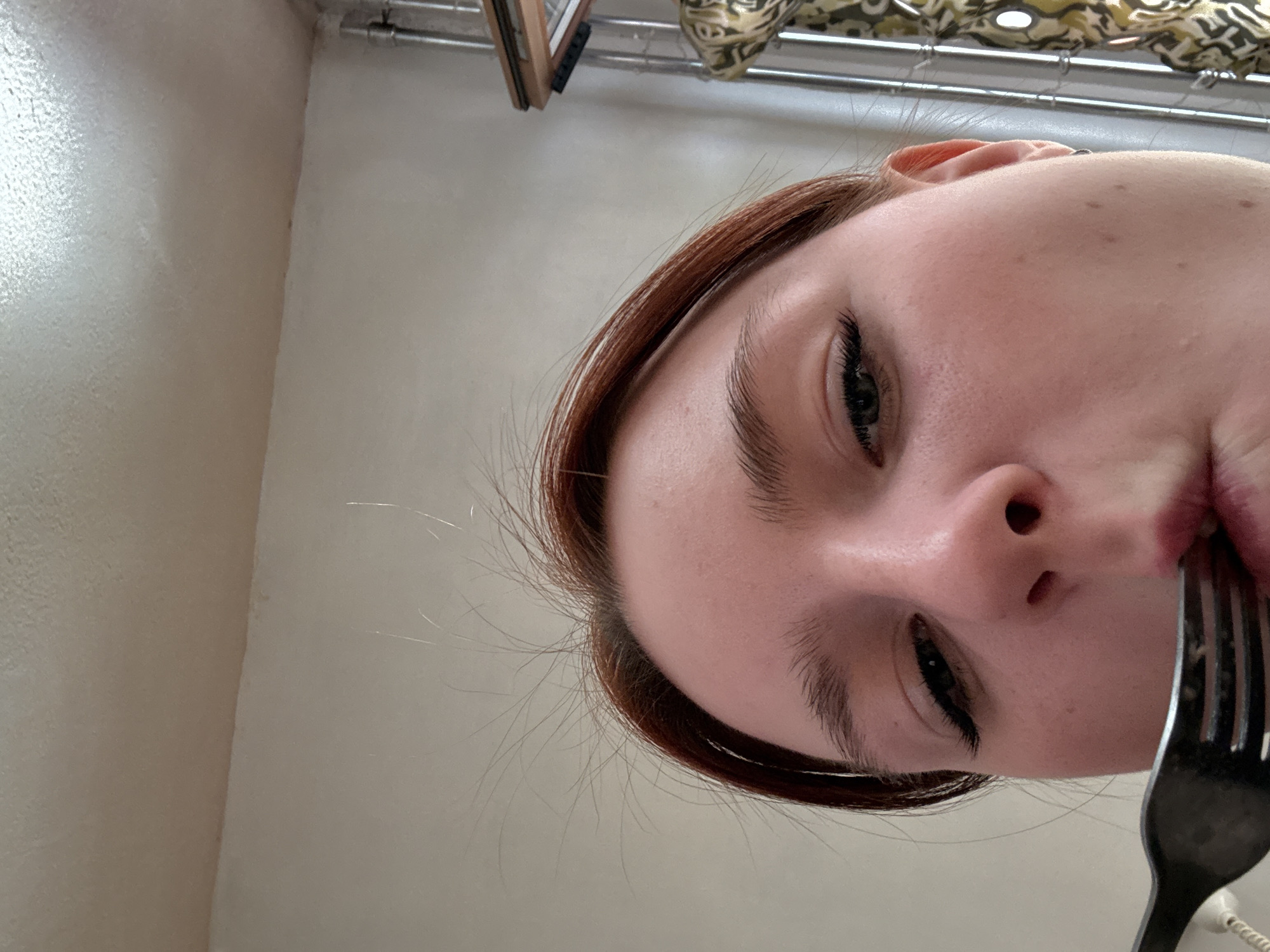 A person's face is centered in the frame, viewed from a low angle looking up towards them. Their face is light-skinned, with noticeable eyelashes and slightly parted lips, with a fork held near their mouth. Their hair is reddish-brown. The background consists of a white wall and a curtain rod near the top edge of the image. The city and location are Unknown.
