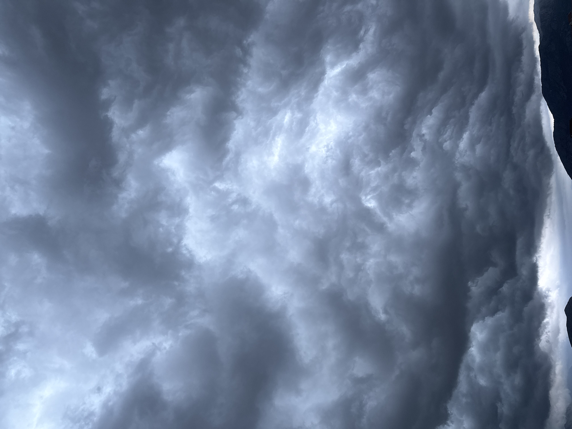 The image displays a dramatic, heavily clouded sky extending across most of the frame, with a dark mountain silhouette visible along the right edge. The sky is dominated by dense, turbulent masses of dark grey and bluish-grey clouds. Patches of brighter, diffused light are visible where the cloud cover is less thick or where light is breaking through, creating a striking contrast and highlighting the voluminous, undulating forms of the clouds.
Towards the right side of the vertically-oriented image, a stark, dark silhouette of a rugged mountain or cliff face is clearly visible, its jagged profile outlined against the brighter, hazy areas of the sky. Some sections of the cloudscape, particularly near the mountainous edge, exhibit distinct wavy or layered patterns, indicating dynamic atmospheric movement.
No people, man-made objects, or visible text are present. The scene evokes a sense of awe and powerful natural forces, suggesting heavy overcast weather, possibly an impending or ongoing storm. The general lighting suggests it could be an overcast day, late afternoon, or early evening. The mountainous terrain is consistent with the provided location context of Podgorica, Montenegro.