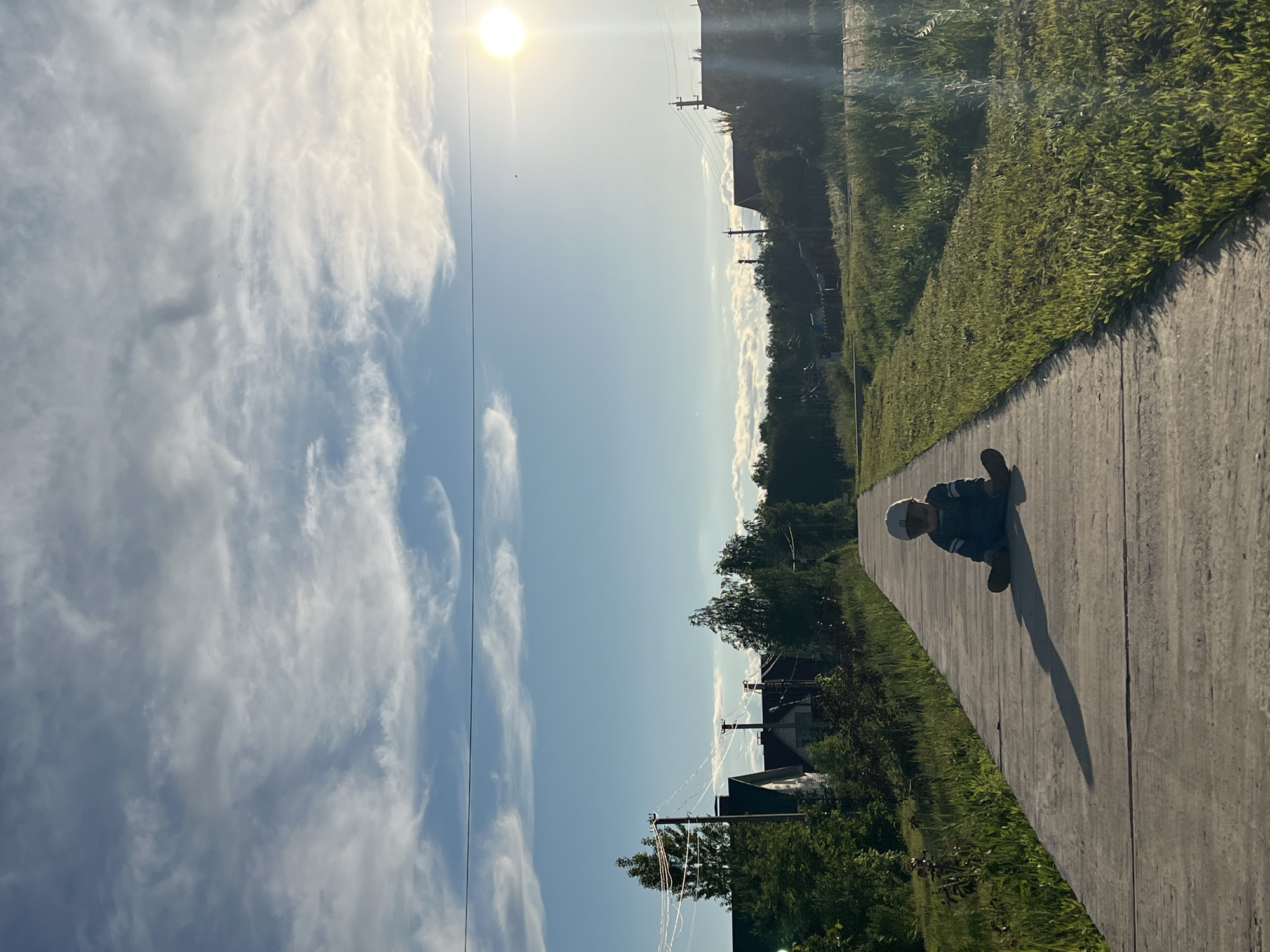 The image, oriented vertically, captures a bright outdoor scene in what appears to be a rural or suburban area, potentially in Noviy, Russia. A young child is positioned on a concrete path, sitting or crouching low, near the right edge of the frame. The child is wearing a light-colored hat and a blue and white striped shirt, with dark shoes. Their body casts a prominent, elongated shadow onto the path, indicating a low sun angle.
The path itself is made of light grey concrete and curves gently. To the left of the path, there is a lush green grassy area with various bushes and trees. Further in the background, a landscape of more trees, residential buildings or houses, and utility poles with overhead wires can be seen, hinting at a settled but open environment.
The sky occupies a significant portion of the image, displaying a bright, clear blue with scattered, wispy white clouds, characteristic of cirrus or altocumulus formations. The sun is visible and intensely bright in the upper right quadrant, suggesting either a morning or late afternoon setting due to its position and the long shadows. The weather appears clear and sunny. No discernible text is visible anywhere in the image.