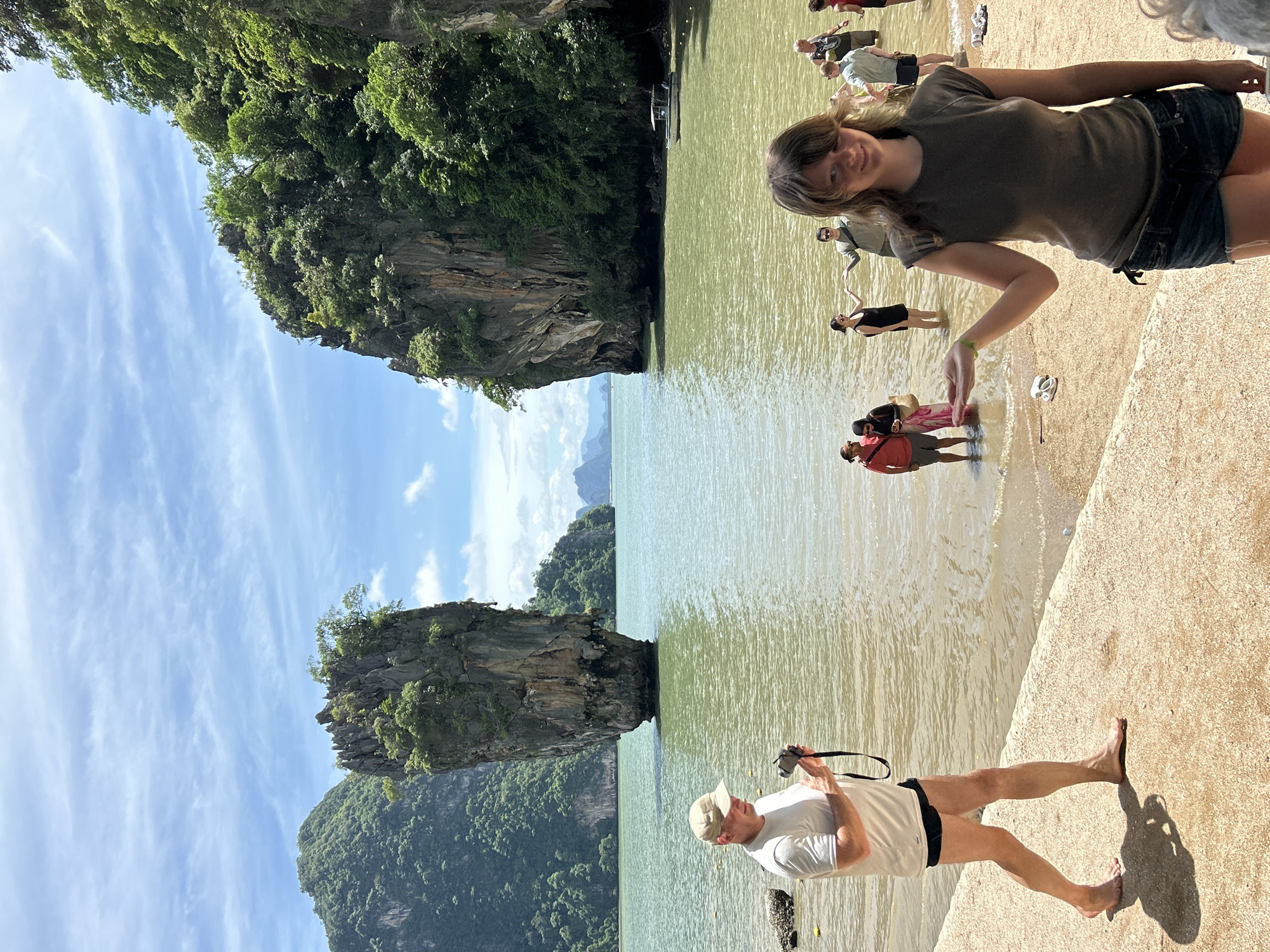 An outdoor, eye-level, medium shot captures a natural landscape dominated by a striking limestone karst formation jutting out of the water, with a lush green jungle covering its peak. The water, a murky green, meets a sandy beach lined with a concrete walkway. In the foreground, a woman with brown hair and bangs, wearing a dark grey t-shirt and denim shorts, smiles at the camera, her right arm extended and palm up. To her left, a barefoot man in a white t-shirt and black shorts, wearing a tan baseball cap, holds a camera. In the background, several people are visible wading in the shallow water. The sky above is a bright blue, dotted with white clouds. The scene is identifiable as Ko Tapu, also known as James Bond Island, located in Phang Nga Bay, near the city of Krasom, Thailand.