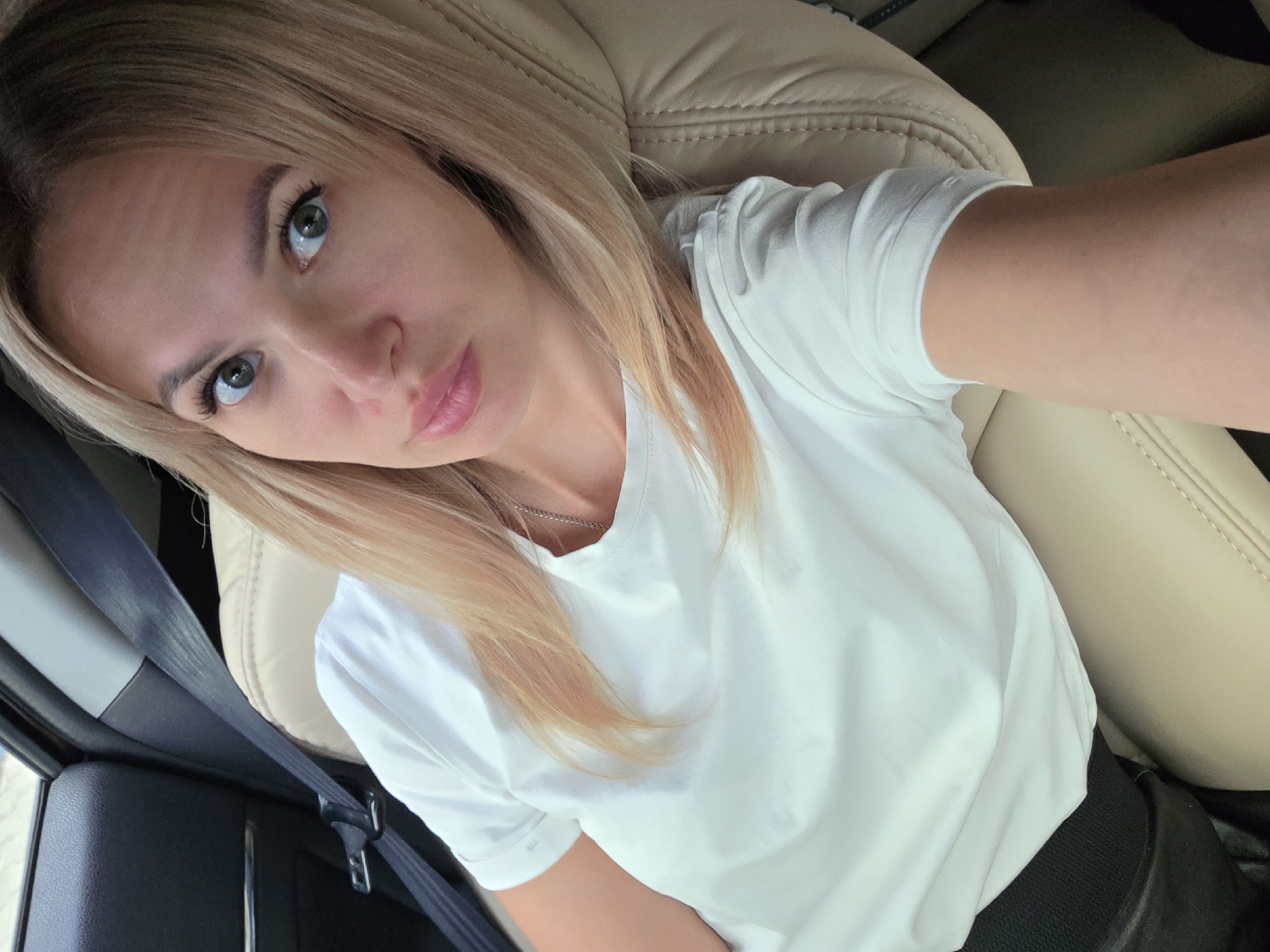 A blonde woman with light skin and blue eyes is sitting in the passenger seat of a car. She is wearing a white t-shirt and a black belt. Her hair is light blonde with some highlights and falls around her shoulders. She is looking at the camera with a slight smile. The interior of the car appears to be leather, with a tan colored seat. A black seatbelt is visible across her shoulder. The background is blurred, suggesting the car is in motion. The image appears to have been taken on a cloudy day, with soft natural light. The location is not specified in the image, but based on the metadata, it is Chaikovsky, Russia.
