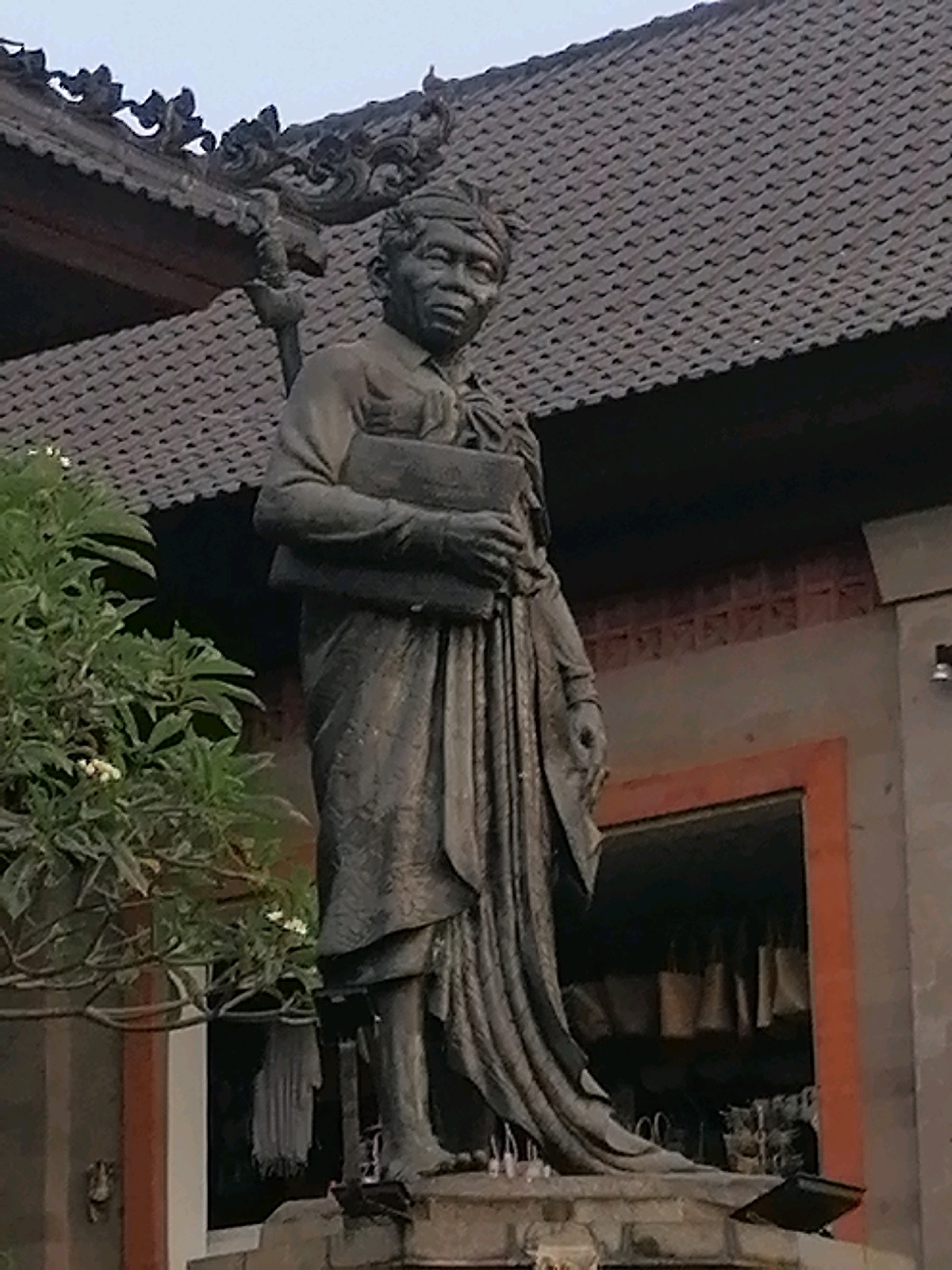 A full-body, slightly blurry, outdoor statue of a man in traditional Balinese attire stands prominently. The statue is dark gray and depicts the man holding a book or scroll in his arms. He is wearing a headcloth and a sarong-like garment draped around his lower body, with a long-sleeved shirt. The statue is positioned in front of a building with a tiled roof and decorative eaves. A lush green tree with small white flowers is visible to the left of the statue. The background shows architectural elements of the building, including a doorway with items hanging inside. The lighting suggests it is either early morning or late evening. The setting appears to be a temple or a public monument in Ubud District, Indonesia.
