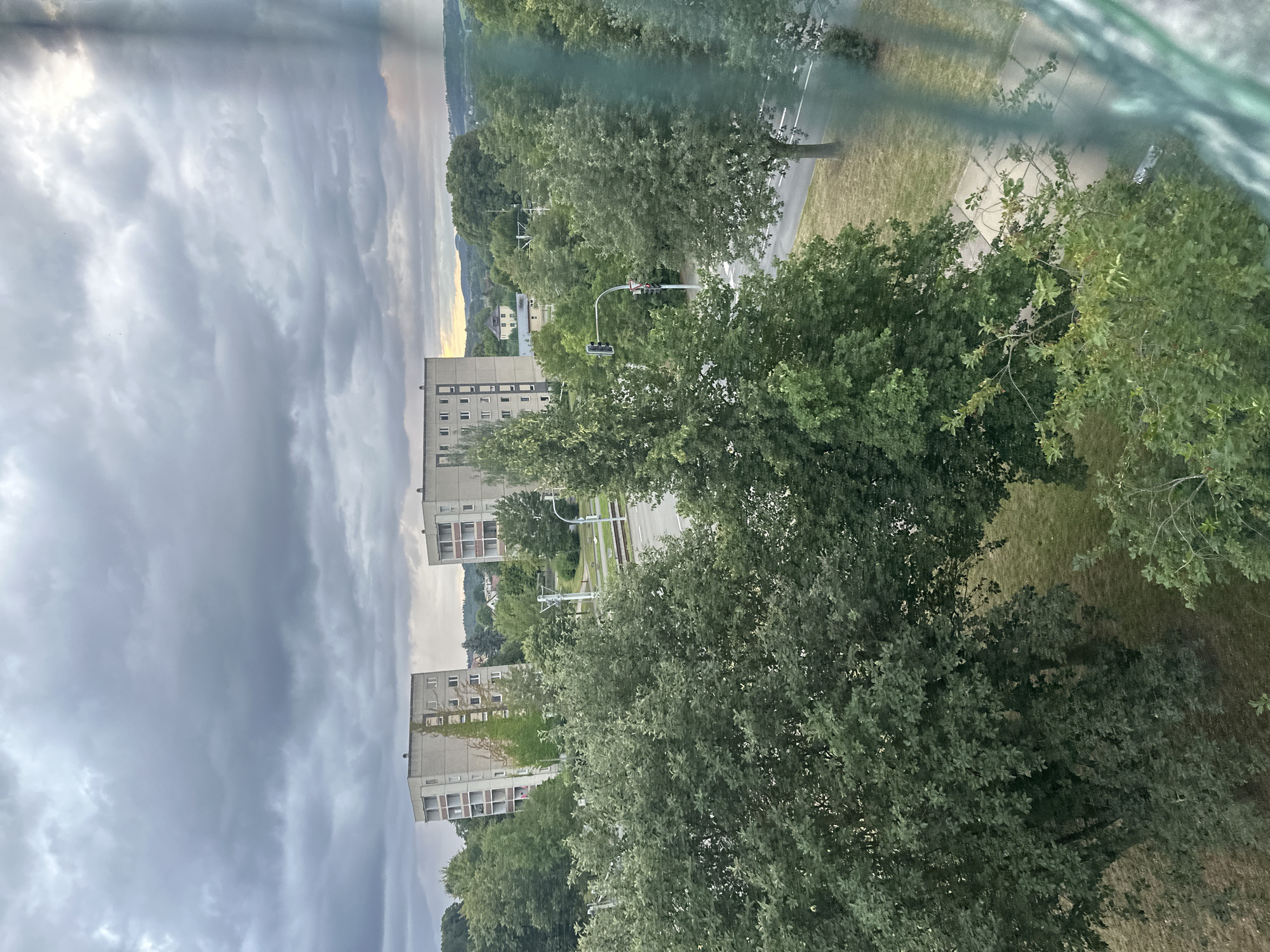 The image captures a view of two tall, grey apartment buildings against a cloudy sky. Green trees dominate the foreground and mid-ground, with some grass and a road visible between them. A traffic light is present on the road, and there are hints of distant hills. The scene appears to be an urban or suburban setting.