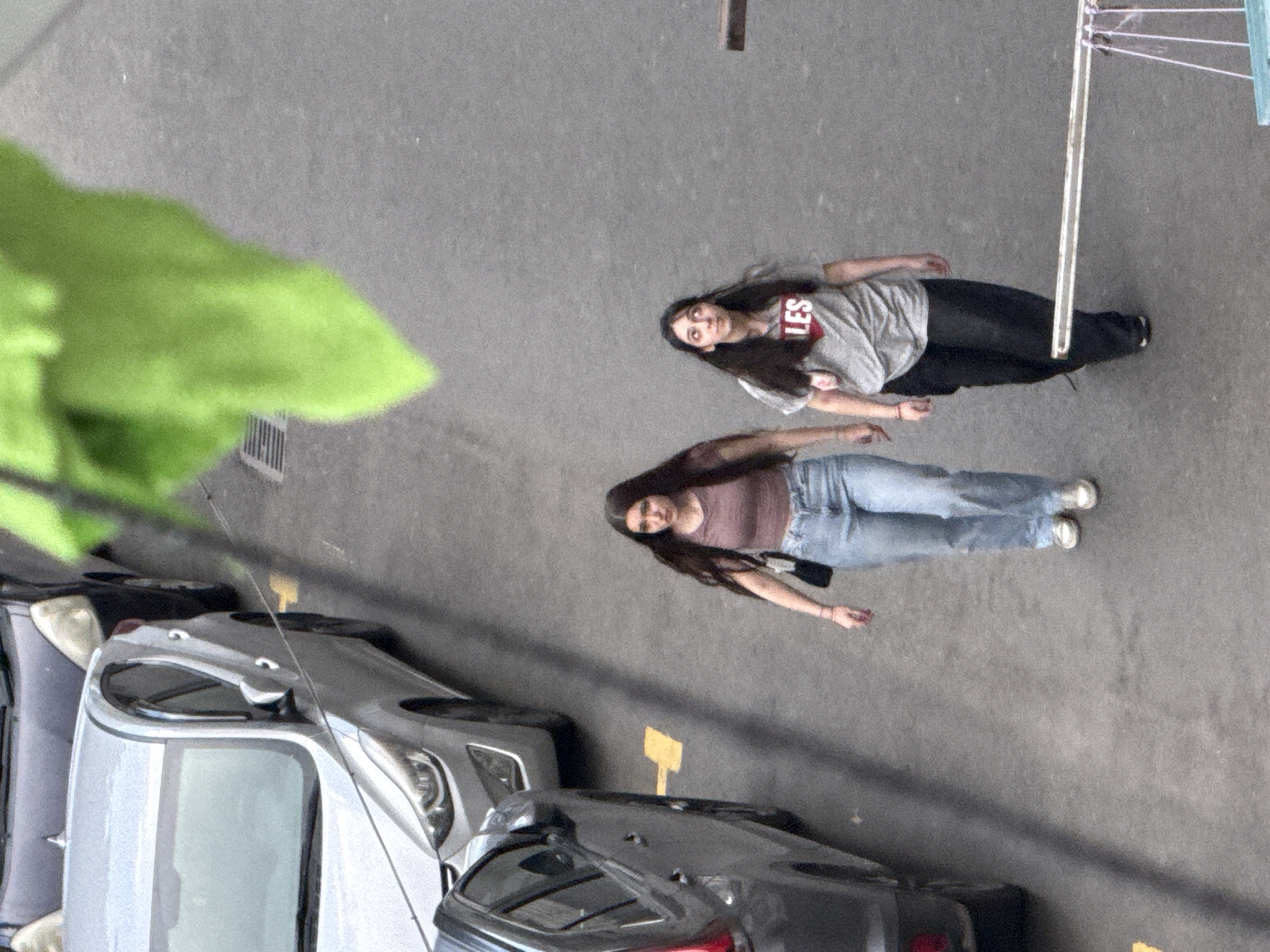The media file depicts an outdoor scene with two young women in a parking lot. The perspective is from above, looking down at the subjects.
The first woman, closer to the top of the frame, is wearing a grey t-shirt with red text that appears to say "LES" in large letters. She is also wearing dark pants and white sneakers. Her hair is dark and long, reaching past her shoulders.
The second woman, positioned below and to the left of the first, is wearing a light brown or pinkish crop top and light blue jeans. She has long, dark hair as well. Her feet are clad in white sneakers. Both women appear to be lying down on the asphalt.
The background shows several parked cars, suggesting an urban or suburban parking area. The ground is concrete or asphalt. A hint of green foliage is visible in the upper left corner, possibly from a plant or tree overhead. In the upper right, a portion of a clothesline or similar structure is visible, with white lines suspended horizontally. Yellow markers are also visible on the ground near the cars.
The lighting suggests it could be daytime, with the sun casting shadows. The overall mood is casual and relaxed, with the women appearing to be posing or resting. There are no explicit indications of the time of day beyond daylight, nor is the weather explicitly discernible, though the lighting might suggest a clear or partly cloudy day. The text "LES" is the only clearly visible text in the image. The location context is Tbilisi, Georgia.