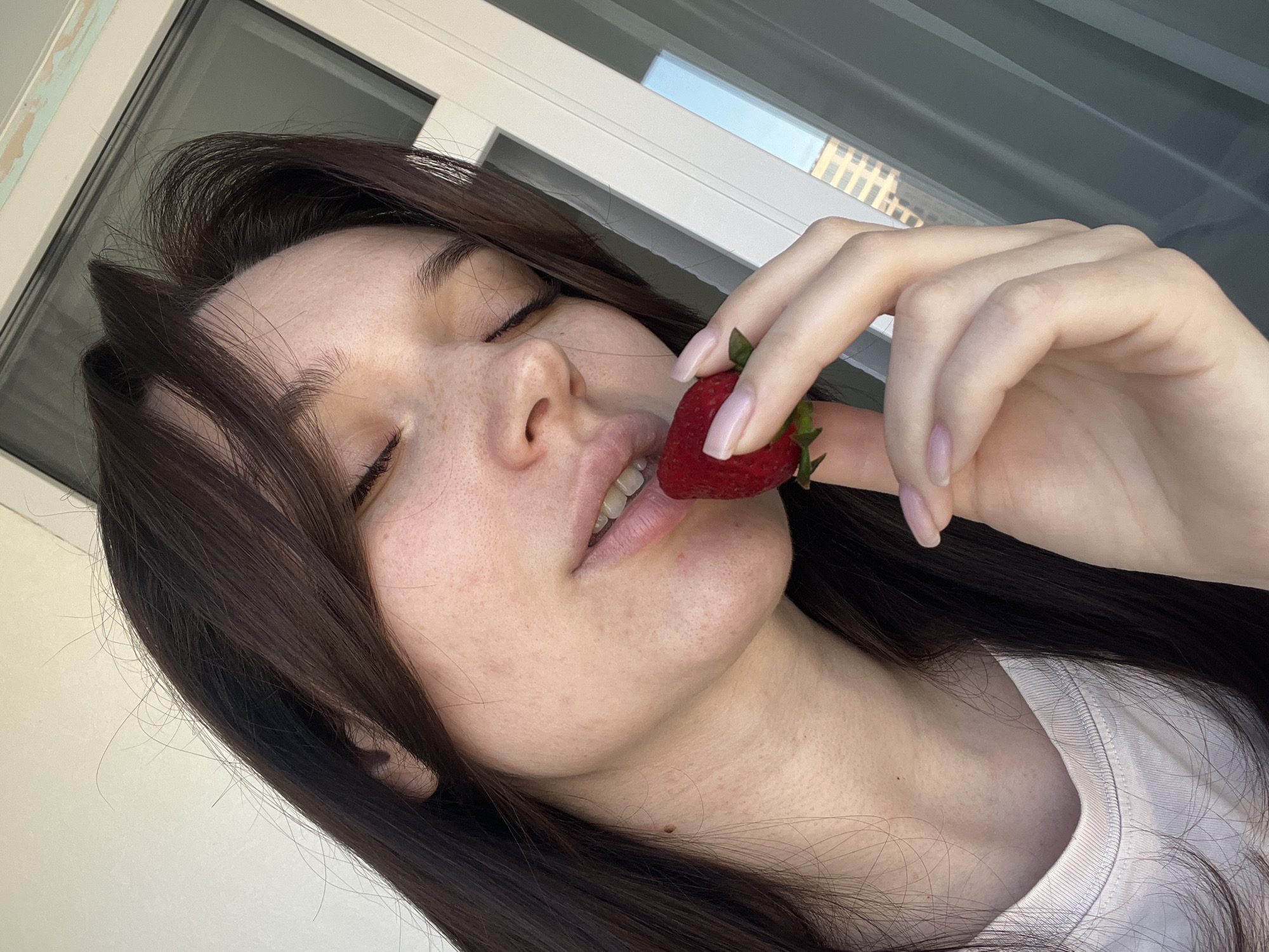 A close-up, angled shot features a young woman in an indoor setting in Tyumen, Russia. Her long, dark brown hair frames her face, with some strands falling across her forehead and shoulders. Her eyes are closed, and her lips are slightly parted, revealing a hint of her teeth, conveying a sense of pleasure or anticipation.
She holds a single, vibrant red strawberry with its green stem and leaves in her right hand, bringing it towards her mouth. Her fingernails are painted a light, natural color. Her fair skin is visible, with some freckles or natural skin texture evident on her forehead and chin. She is wearing a simple white t-shirt, visible on her right shoulder and chest.
The background consists of a large window with white frames, indicating an interior space with ample natural light. Through the window, reflections of a modern building with multiple windows are discernible, suggesting an urban environment. The bright natural light indicates it is daytime. There is no visible text in the image. The composition creates an intimate and serene atmosphere focused on the woman and the fruit.