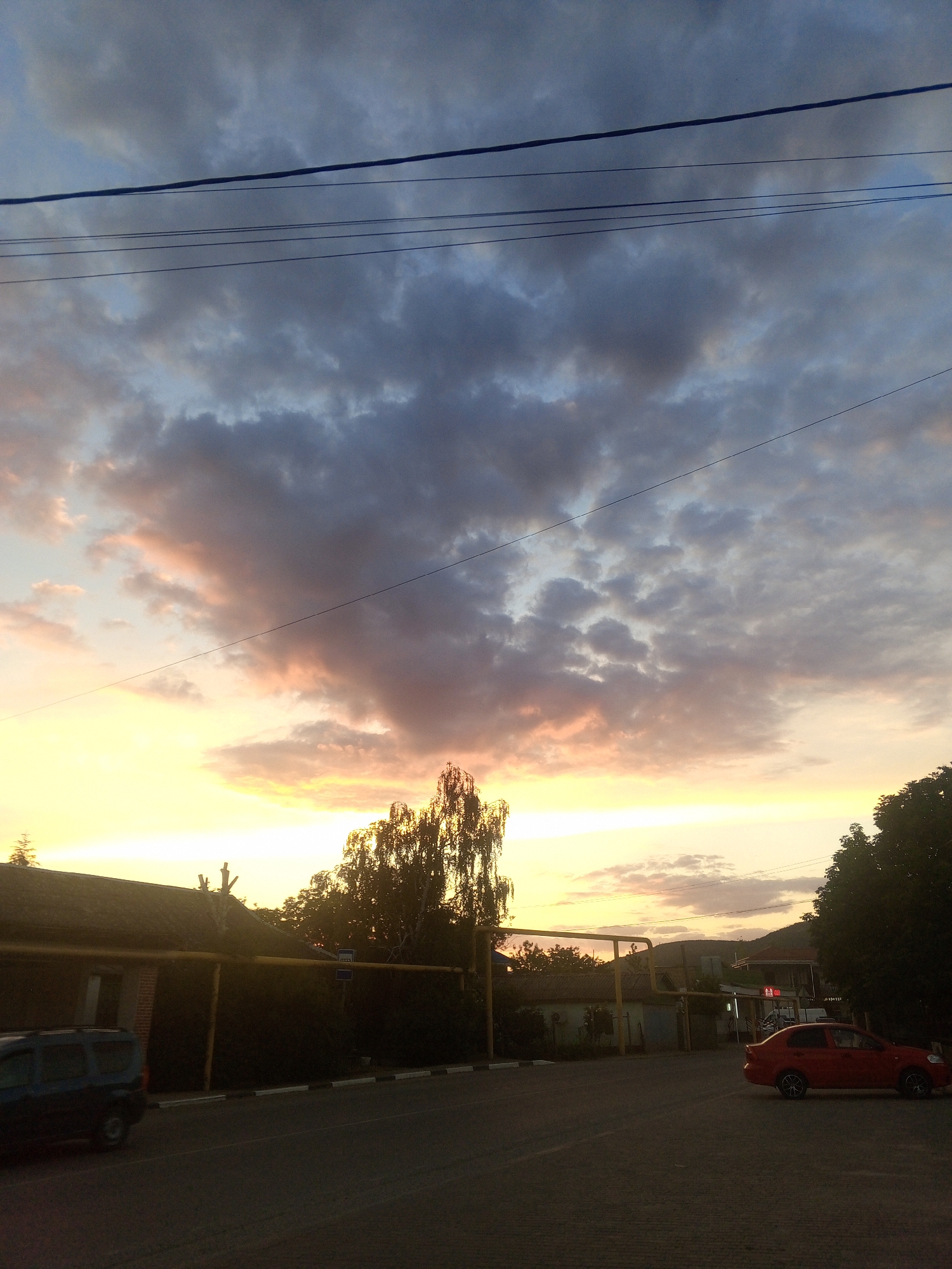 This is a vertical, low-angle shot of a street scene at dusk in Голубинское сельское поселение, Ukraine. The sky is filled with dramatic, colorful clouds, ranging from dark gray to soft pink and bright yellow near the horizon. Several power lines are strung across the upper portion of the frame. In the foreground, a dark asphalt road is visible. To the left, a dark blue minivan is parked. In the middle ground, a low-rise building with a dark, sloped roof is partially obscured by trees and a yellow pipe running horizontally in front of it. To the right, a red sedan is parked on the asphalt. A tall, slender tree with dense foliage is positioned between the building and the red car. In the background, more buildings, trees, and a hillside can be seen under the twilight sky.