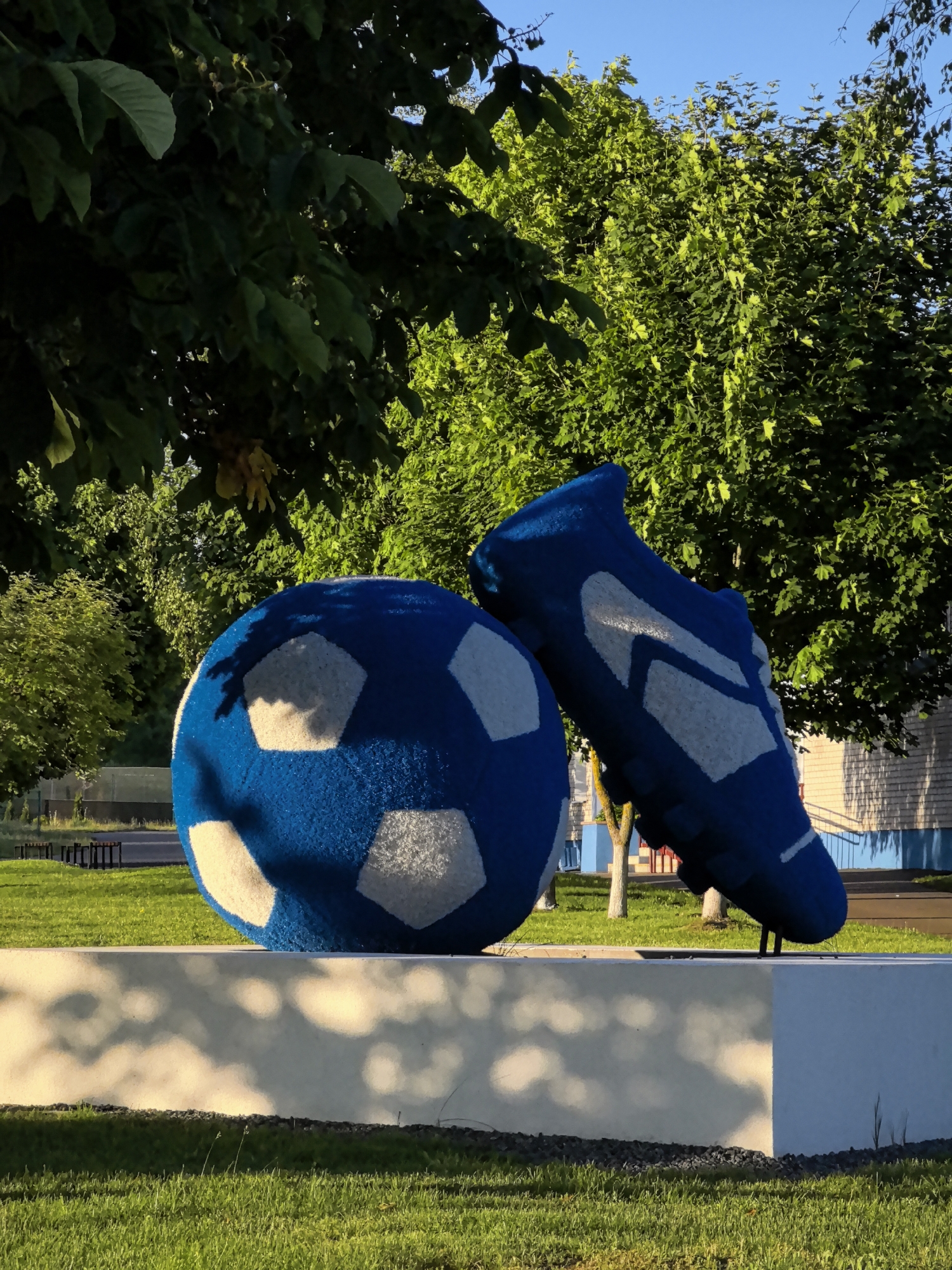 A large, blue soccer ball and a soccer cleat are displayed on a white pedestal outdoors. The soccer ball is positioned to the left and is round with white hexagonal panels. The soccer cleat is on the right, upright and appearing to be a replica of a soccer shoe with white detailing. Both are brightly colored and appear to be made of a fuzzy material. Behind the pedestal, a patch of green grass is visible, and beyond that, trees with lush green leaves fill the background. The sky above is clear and blue. The scene is illuminated by natural sunlight, casting shadows on the grass and pedestal.