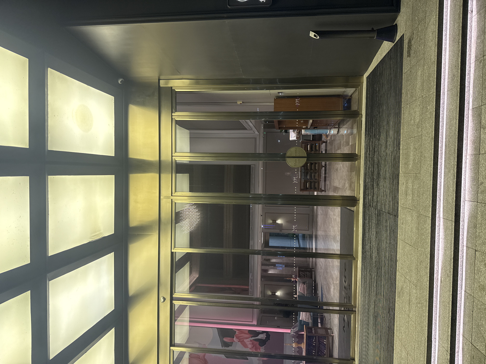 The image shows the entrance to the Mercure Hotel in Huangpu District, China, at night. The entrance features large glass sliding doors with gold-colored frames. Above the entrance, a ceiling with translucent panels is illuminated from within. A security camera is visible on the ceiling near the right side of the entrance.
Inside the hotel, visible through the glass doors, is a well-lit lobby with a marble floor. There are seating areas with sofas and tables. A reception desk or counter is also partially visible. In front of the glass doors, there is a black ribbed doormat. The steps leading up to the entrance are made of stone and are illuminated with white lights.
To the right of the entrance, a dark gray wall is visible, with a tall, slender black object that appears to be an outdoor ashtray or waste receptacle. A small white security camera is mounted on the wall above the entrance. Another sign with a logo and text is partially visible on the far right edge of the image. The overall scene suggests a modern, upscale hotel entrance.