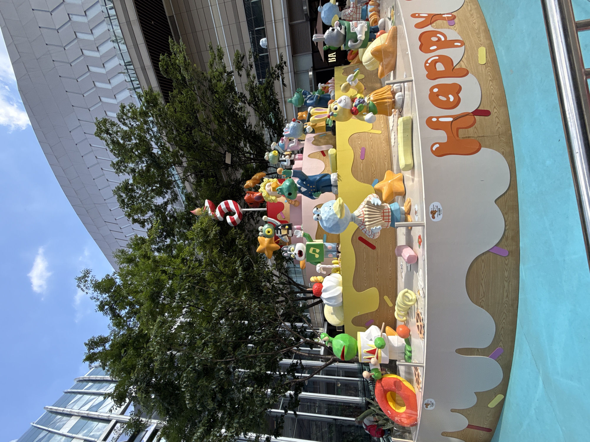 This outdoor shot depicts a multi-layered display of cartoon character figures arranged on a structure resembling a cake, with the word "Happy" prominently displayed. The scene is set in an urban environment, as evidenced by the modern architectural building in the background. Green trees partially obscure the building. A blue sky is visible in the upper left corner. The precise location is Liangjiang, China.