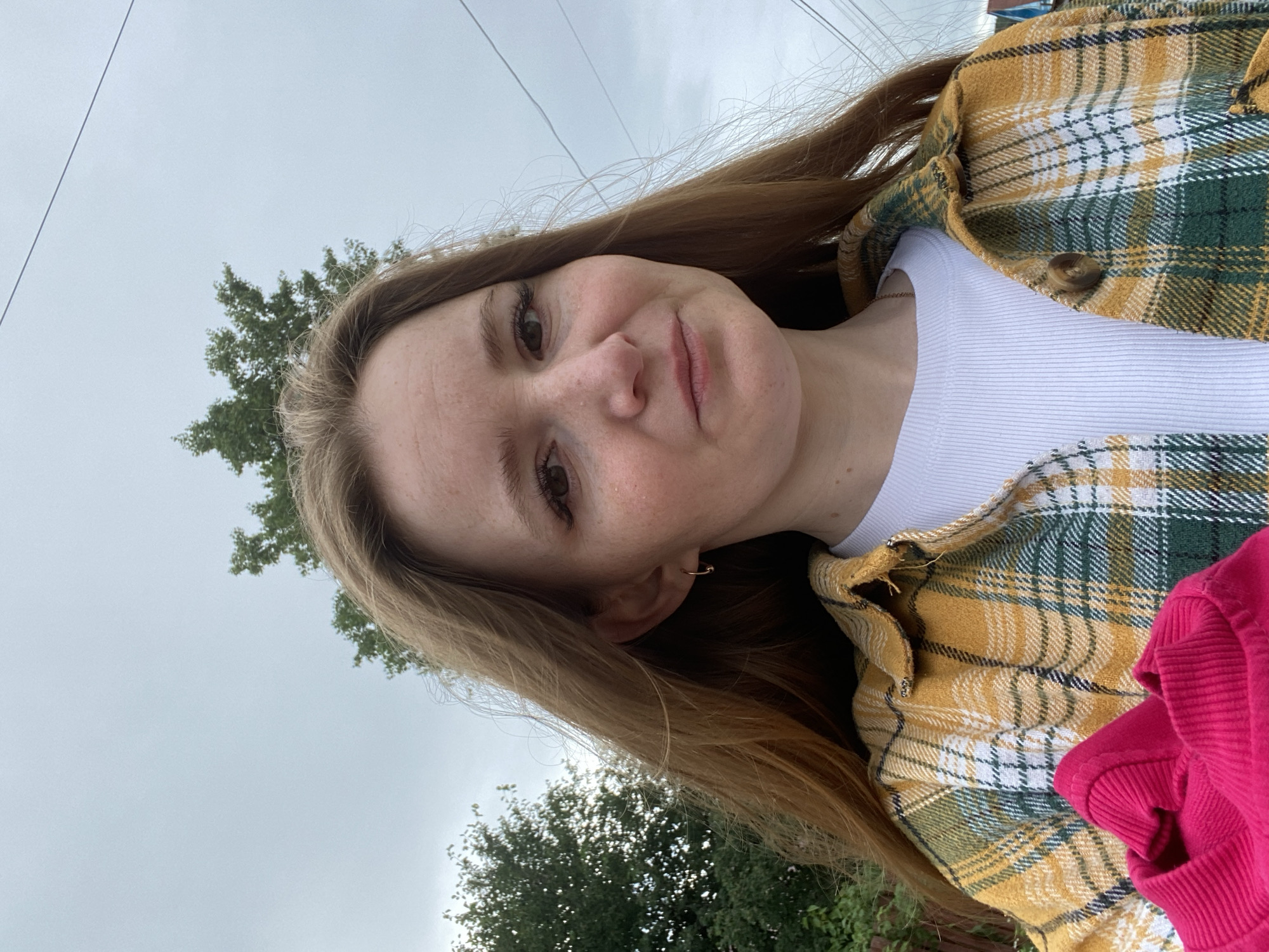 A young woman with long, auburn hair is taking a selfie outdoors. She is wearing a white ribbed t-shirt under an open, yellow and green plaid flannel shirt. A bright pink garment is held in front of her chest. Her facial features include light skin with freckles, brown eyes, and light makeup. The background shows a cloudy sky, green foliage from trees, and utility wires. The image appears to have been taken in a semi-rural or suburban area.