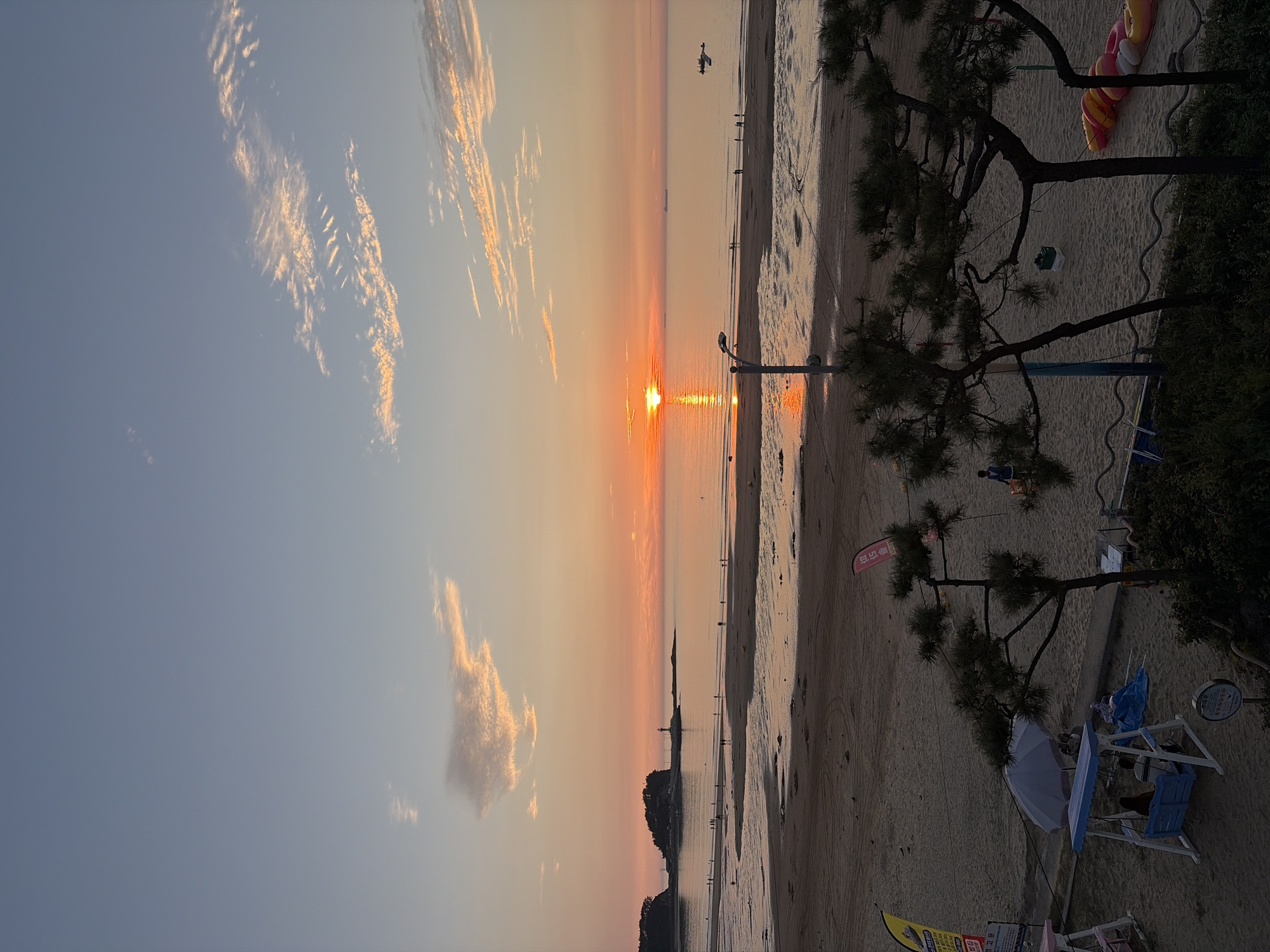 The image captures a sunset scene at Mohang Beach in Mohang-ri, South Korea. The sky transitions from a pale blue at the top to shades of orange and pink near the horizon, with scattered, wispy clouds illuminated by the setting sun. The sun, a bright orange orb, is partially submerged on the horizon, casting a shimmering reflection on the calm sea.
The beach in the foreground is sandy with tidal pools reflecting the sky. Several people are visible as small figures strolling along the waterline and on the wet sand. In the middle ground, a body of water stretches out to the horizon. On the left, a small island with trees and a lighthouse can be seen. Farther out in the water, several ships are present.
In the immediate foreground, the beach transitions to a grassy area with trees, some of which are pine trees with dark green needles. A lamppost stands on the beach. Further into the foreground, there are recreational elements like a blue and white swing set with two people sitting on it, an umbrella providing shade, and some inflatable water toys arranged on the sand. Two flags, one yellow and one pink, are also visible on the beach. The overall atmosphere is serene and tranquil, characteristic of a beach at dusk.