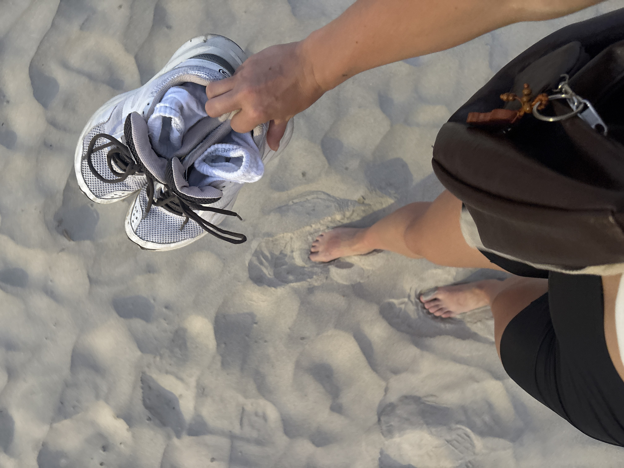 The image is a high-angle, top-down shot capturing a person's hand, feet, and lower body on a sandy surface, likely a beach in Đà Nẵng, Vietnam. The person is holding a pair of grey and white athletic shoes with dark laces, which have white socks stuffed inside them. Their right hand firmly grips the shoes.
The person's bare feet are visible on the light-colored sand, with some footprints already imprinted, suggesting they have been walking or standing in the sand. Part of their lower body is visible, dressed in black shorts and a dark top or bag strap that covers their shoulder. A dark shoulder bag or backpack is visible on the right side of the frame, from which a keyring with a small, ornate cross charm and another small charm hangs from a zipper pull.
The sandy ground appears slightly undulating with shallow depressions and foot impressions. The lighting suggests it is daytime, with soft shadows indicating a possibly overcast sky or diffused light, creating a calm atmosphere. The overall scene conveys a sense of leisure or activity at a beach, where one might remove shoes to enjoy the sand. The only discernible text is a partial logo "C-T" on the side of one of the shoes.