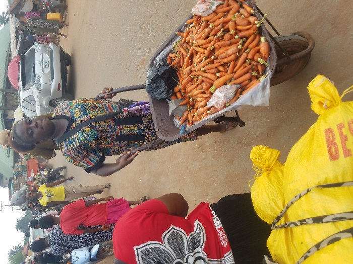 The image depicts a busy street market scene in Nnobi, Nigeria, during daylight hours.
In the midground, a dark-skinned woman with dark hair pulled back, wearing a multi-colored patterned short-sleeved tunic and sandals, is actively pushing a brown wheelbarrow. The wheelbarrow is filled with numerous orange carrots, some loose and some contained in small, clear plastic bags. A black plastic bag is also visible within the wheelbarrow. The woman has a dark bag with a purple and white patterned strap slung across her body.
In the immediate foreground, to the bottom left, several large yellow sacks are visible, partially secured with striped fabric. Red text, including "BEG," is legible on one sack. Behind these sacks, the upper back of a person wearing a red shirt with a white floral pattern is partially visible, facing away from the viewer.
The background reveals a bustling unpaved road. On the left, a light blue scooter is parked. Several pedestrians are walking, including a person in a red t-shirt and pink skirt, another in a black and white patterned dress, and a woman in a yellow t-shirt and light brown pants carrying an object on her head, walking towards the viewer. A man on a motorcycle is further back on the road. Buildings with diverse roofing materials, including corrugated iron, line the street.
On the right side of the background, a silver SUV, appearing to be a Lexus RX model, is either parked or moving slowly. Additional pedestrians are present, one carrying a yellow jerrycan. A red umbrella-like structure is visible above market stalls. The ground throughout the visible street area is dusty, light brown earth. Overhead, power lines are discernible against the sky.