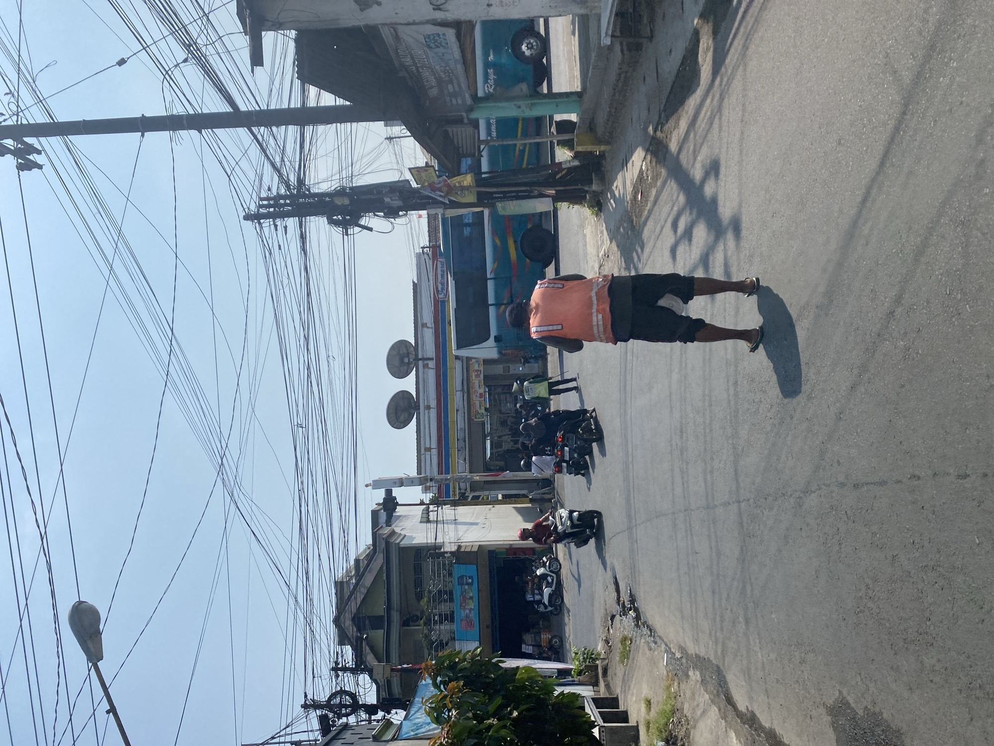 The image shows a street scene in Kediri City, Indonesia. In the foreground, a person wearing an orange safety vest and dark shorts walks away from the camera. They are wearing flip-flops. The street is paved and shows some wear and tear, with a gutter on the left side. Various motorcycles are parked and in motion on the street.
On the right side of the street, there is a building with a small shop or kiosk. A utility pole with a dense network of overhead wires stands prominently. Further down the street, a blue and white bus with colorful stripes is visible, along with a building that appears to be a convenience store, possibly an Indomaret, indicated by its signage. The sky is clear and bright, suggesting a sunny day. Buildings lining the street appear to be a mix of commercial and residential structures, with some advertising signs visible. The overall impression is a bustling urban street with active transportation and commerce.