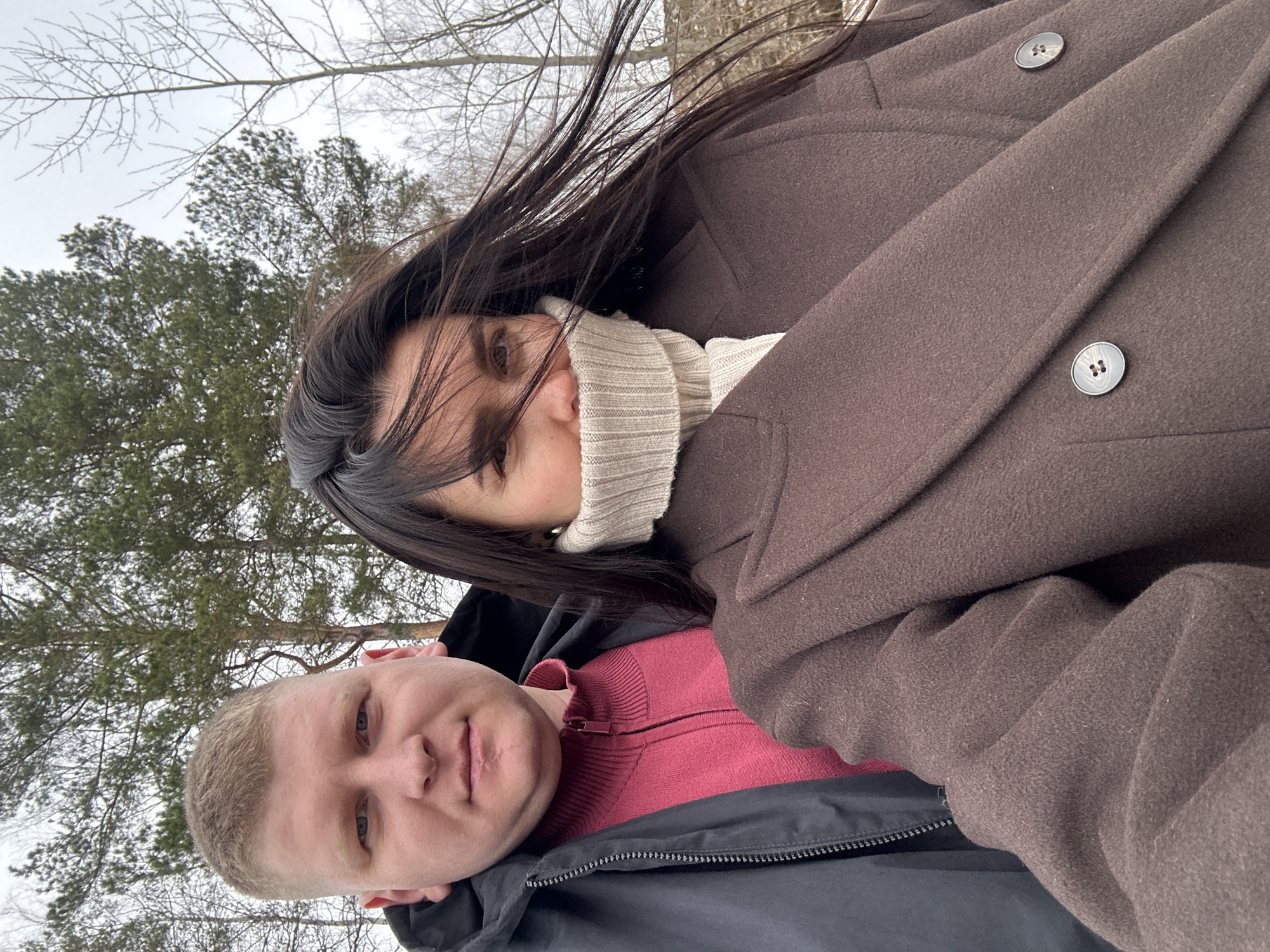 A vertically oriented photo captures two individuals, a woman and a man, taking a close-up selfie outdoors in what appears to be a park or forest in Chaikovsky, Russia. The scene is set against a backdrop of bare deciduous trees and green evergreen trees under an overcast, light grey sky, suggesting a cold season like late autumn, winter, or early spring.
The woman, positioned higher and partially overlapping the man, has long, dark hair that appears to be blowing to her left. She wears a light-colored (likely beige or off-white) ribbed turtleneck sweater and a dark brown, heavy coat with visible large, dark buttons. Her gaze is directed towards the camera, and her expression is neutral to slightly soft.
Below and to her right, the man has short, light-colored hair and light eyes. He is wearing a reddish-pink zippered sweater underneath a dark (possibly black) jacket. He also looks directly at the camera with a neutral expression.
Their close proximity indicates intimacy or camaraderie. The clothing suggests a chilly outdoor temperature. No visible text is present in the image.