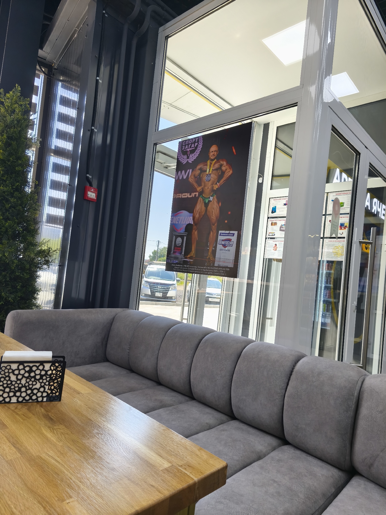 The image depicts the interior of a modern establishment with large windows, likely a cafe or lounge, located in Komsomolsk-on-Amur, Russia. In the foreground, a polished wooden table with a black napkin holder containing white napkins is visible. Behind the table, a large, plush grey sectional sofa occupies the lower portion of the frame. The background features a large window showcasing a poster of a muscular bodybuilder wearing a medal and swim briefs. Text on the poster appears to be in Russian, with visible words like "СОРТ" (Sport) and a year. Through the window, a silver van is parked on a street, suggesting an outdoor environment with trees and buildings visible in the distance. The overall scene is well-lit, with natural light streaming in from the windows. The establishment appears to be clean and well-maintained.
