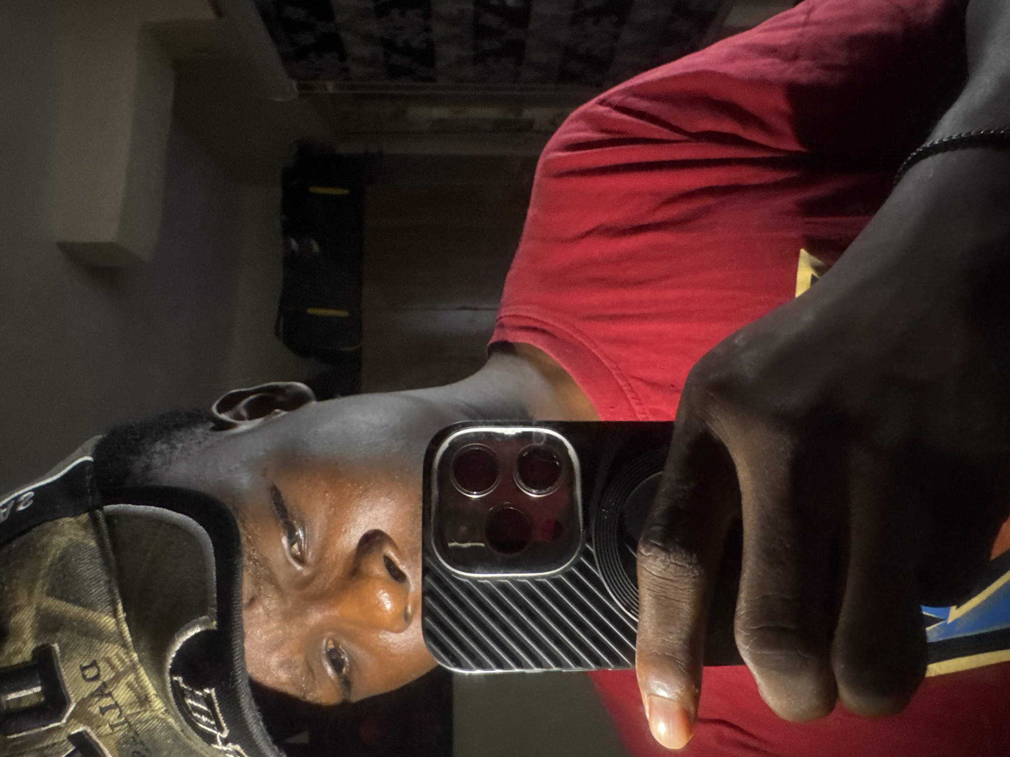An image, appearing to be a reflection or mirror selfie, depicts a dark-skinned individual indoors in Ihiagwa, Nigeria. The subject, likely male, is shown from the chest up, wearing a red t-shirt and a camouflage-patterned baseball cap. They are holding a smartphone in their right hand, with the index finger extended. The phone's three-lens camera array and a distinctive textured, striped case are prominently visible. A black beaded bracelet adorns the person's left wrist. The individual's face is visible, looking towards the camera with a neutral expression.
The background is dimly lit, showcasing a light-colored wall or shelf on the left and a dark, rectangular object, possibly furniture, further back. A patterned surface, suggestive of a ceiling or wallpaper, is also present. Visible text on the baseball cap includes "DYNASTY" or a similar word on the front, and "SA" or "3A" on the side of the brim. A partial yellow and blue graphic design is discernable on the red t-shirt. The overall low-light conditions indicate an indoor setting, potentially during the evening or in a dimly lit room.
