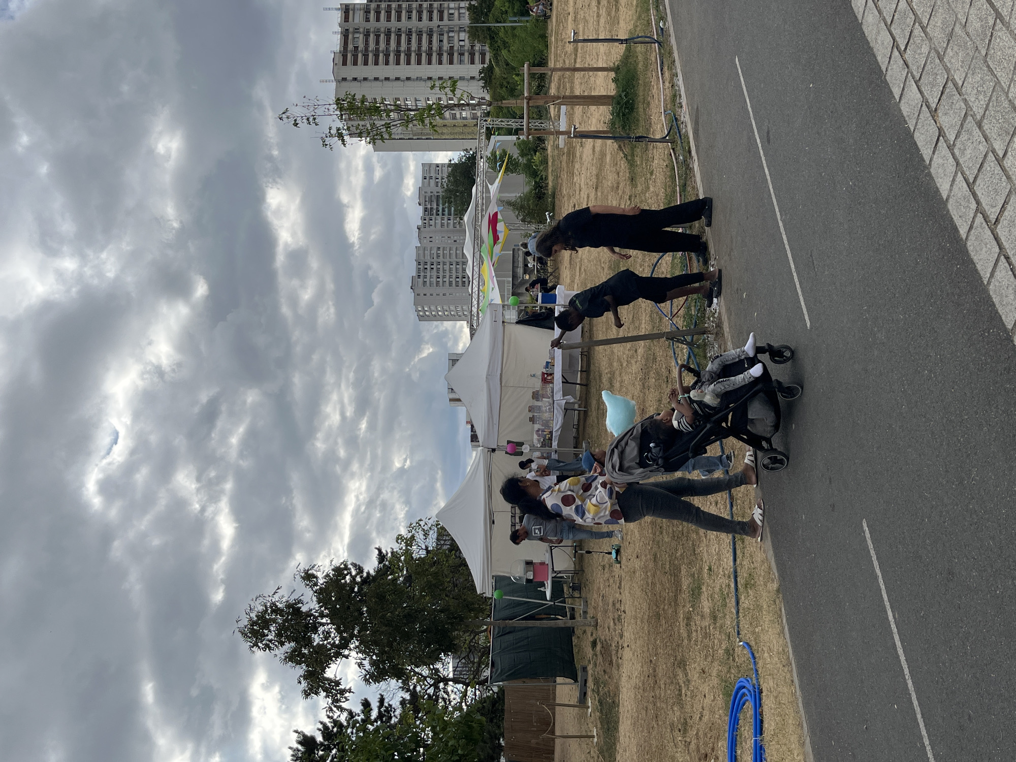 An outdoor scene in Bobigny, France, features a cloudy sky over a dry, grassy field with several white tents. In the foreground, a paved pathway runs along the bottom of the image. To the left, a blue hose is coiled on the ground. Several people are present. A woman in a colorful shirt pushes a stroller containing a young child. Two individuals, one appearing to be an adult male and the other a young male, are standing near a metal structure. Another adult male and female are observed near the tents. In the background, trees and tall apartment buildings are visible. One tent displays a variety of colorful items on tables.