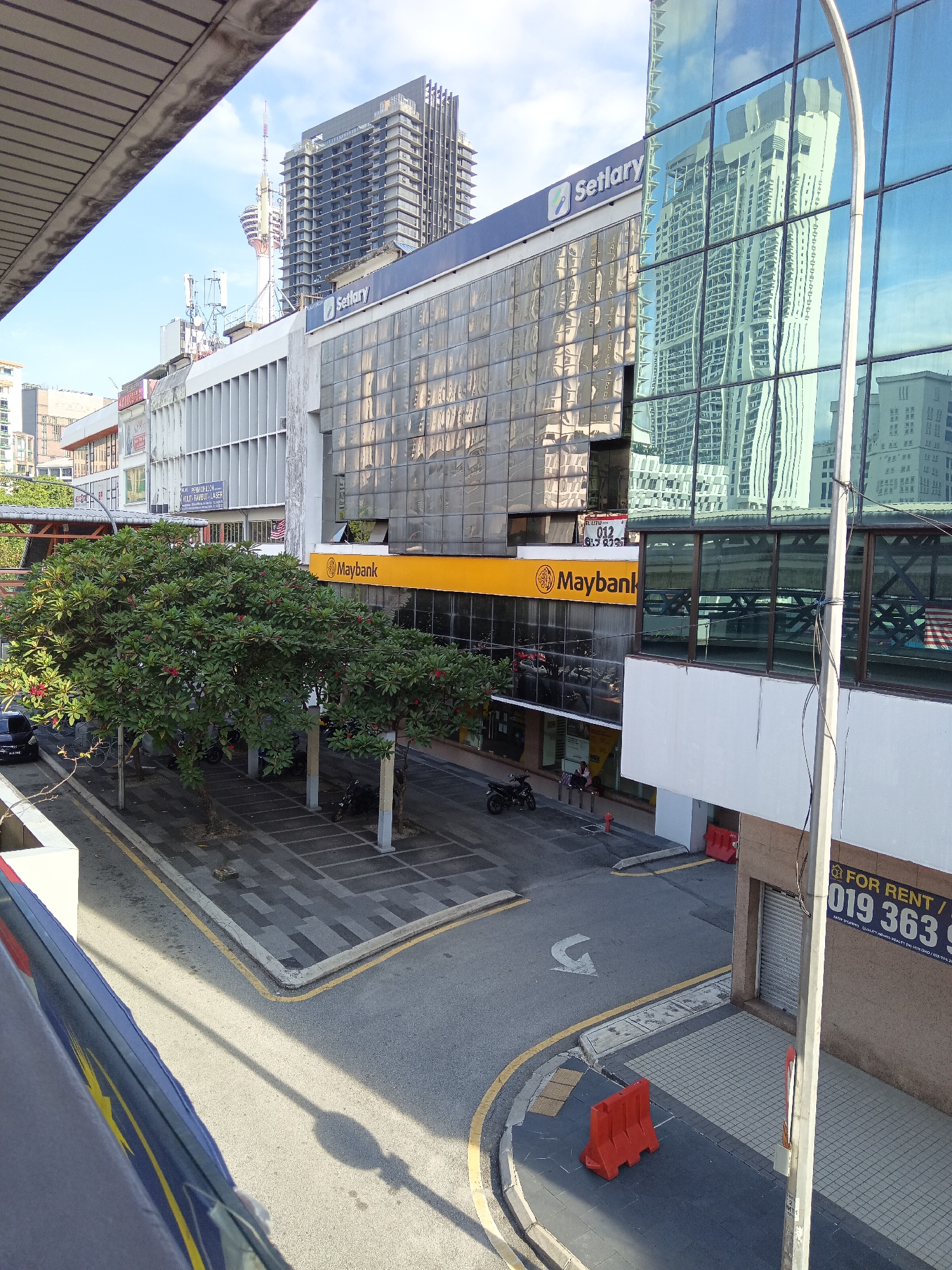 The image depicts a street scene in Kuala Lumpur, Malaysia, dominated by the architecture of urban commercial buildings. In the foreground, a grey overhead walkway structure partially obscures the view. Below, a street with a grey asphalt surface is marked with yellow lines and a white arrow indicating traffic flow. To the right, a sidewalk area with a checkered tile pattern is visible, bordered by a curb. A tall, grey utility pole stands prominently on the sidewalk.
A prominent yellow awning with the "Maybank" logo runs across the facade of a building. Above the awning, a glass-fronted building displays reflections of other skyscrapers, characteristic of Kuala Lumpur's modern skyline. A sign reading "Setlary" is also visible on this building. Further back, a tall, modern skyscraper with a dark facade and a distinctive top structure is a significant feature of the background, along with the Kuala Lumpur Tower in the distance.
Several trees with lush green foliage and red flowers are planted along the sidewalk, providing a touch of nature to the urban setting. Parked vehicles, including a black car and several motorcycles, are visible in the street and on the sidewalk. Two individuals are seated on a bench near the Maybank entrance, and two red traffic barriers are placed on the sidewalk. A sign with "FOR RENT" and a phone number is attached to a utility pole. The sky above is clear with a few wispy clouds, and sunlight casts shadows across the scene.