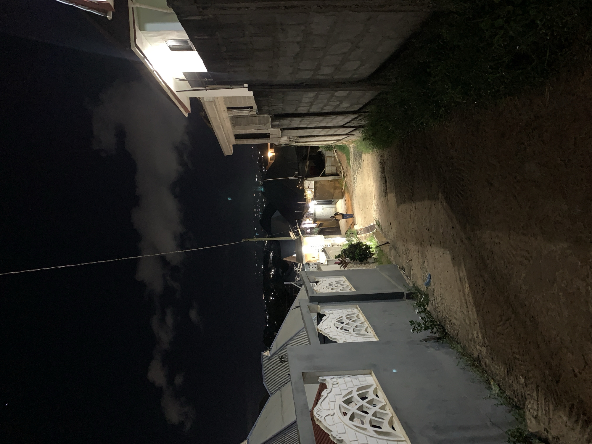 The image shows a narrow alleyway at night in Dar es-Salaam, Tanzania. The path is unpaved and illuminated by dim, distant lights. On one side, there is a tall, unfinished concrete wall. On the other side, there is a multi-story building with decorative window frames. A person wearing dark clothing is walking towards the end of the alley. The sky is dark, with some cloud cover visible in the distance. City lights are scattered in the background.