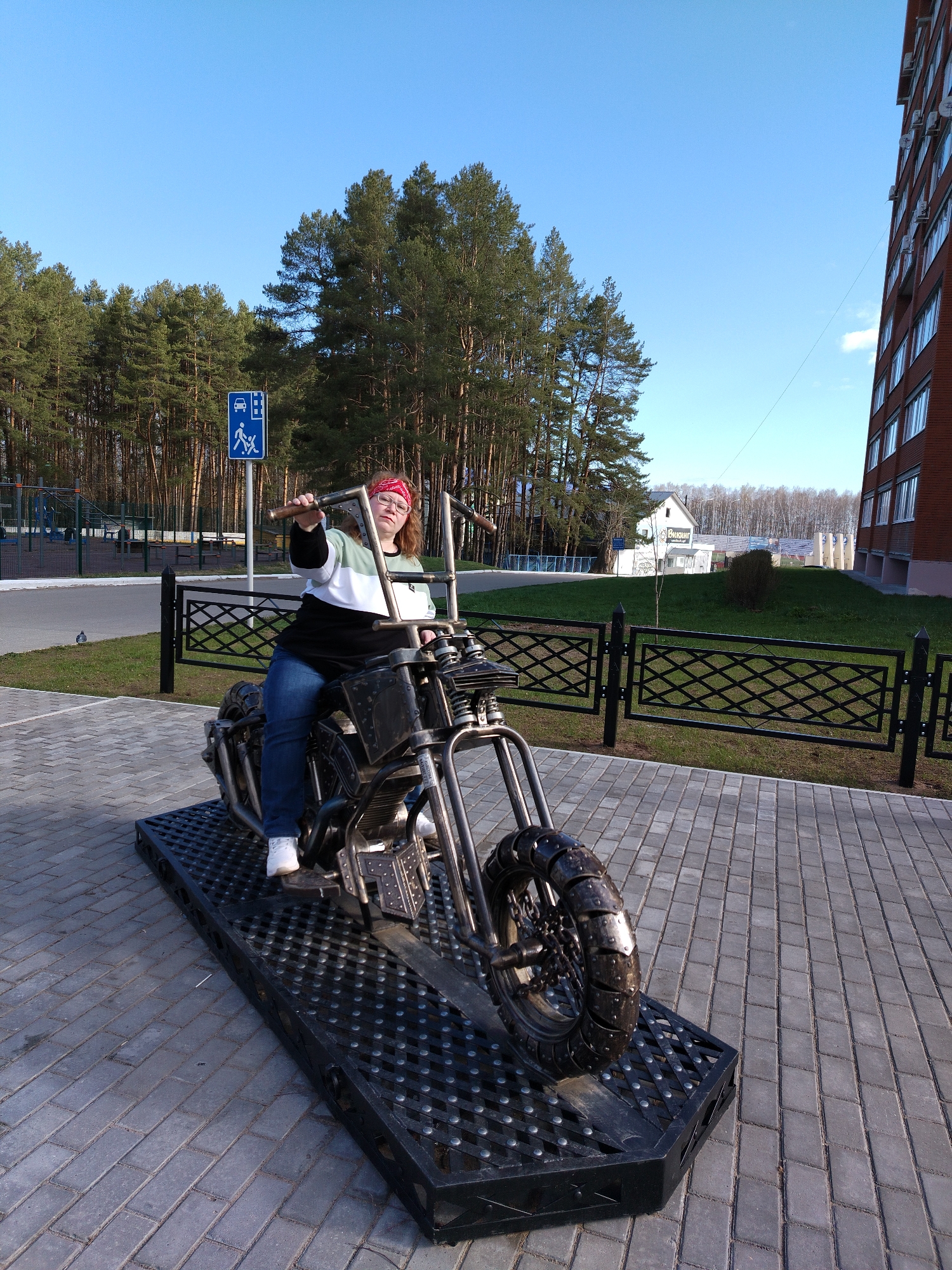 A fair-skinned female with reddish hair sits atop a metal sculpture of a motorcycle. She wears a red bandana, glasses, a sweater with white, green, and black stripes, blue jeans, and white sneakers. She grips the motorcycle handlebars with her right hand extended outward. The motorcycle sculpture rests on a black metal grate on a paved surface of gray bricks. A black metal fence borders a grassy area with a small sapling and a building in the background. Evergreen trees are visible in the background under a blue sky. A sign indicating a parking area for vehicles with disabilities is present on the left. In the background on the right, a brick building with multiple windows is visible. The location is in the city of Chaikovsky, Russia.
