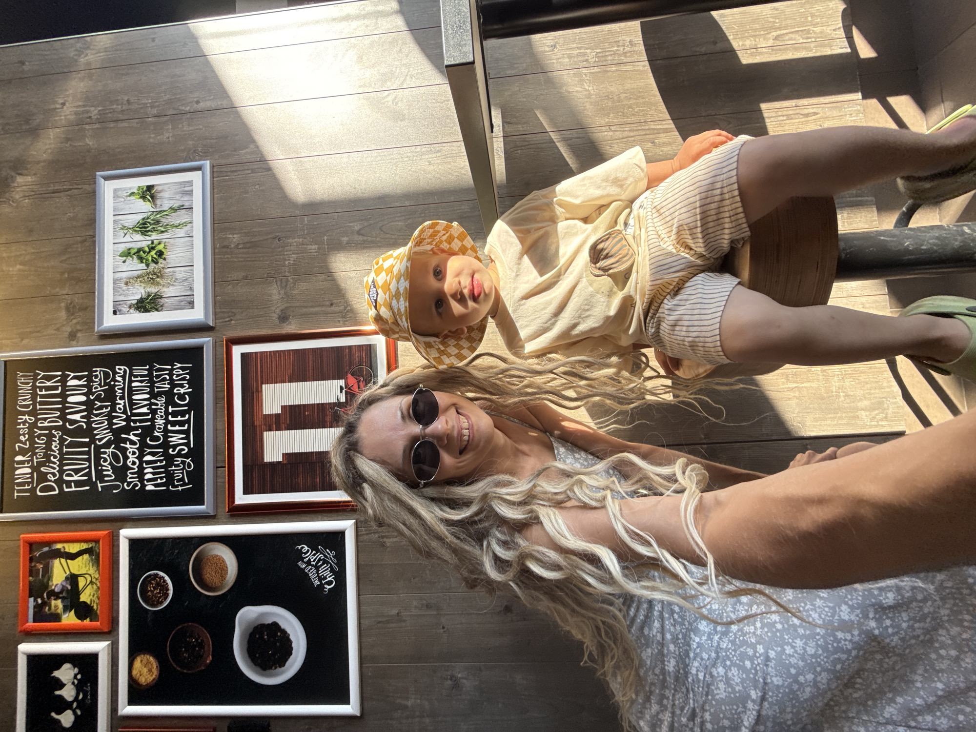 In an indoor setting with wood-paneled walls, a woman with long blonde dreadlocks and sunglasses smiles at the camera, holding a selfie stick. Beside her, a young child wearing a checkered hat and a light-colored t-shirt sits on a stool. The background features several framed pictures on the wall, including one with text and another with an illustration of the number 11. The lighting suggests it is daytime, with sunlight casting shadows. The overall scene appears to be a casual, possibly a cafe or restaurant, environment.