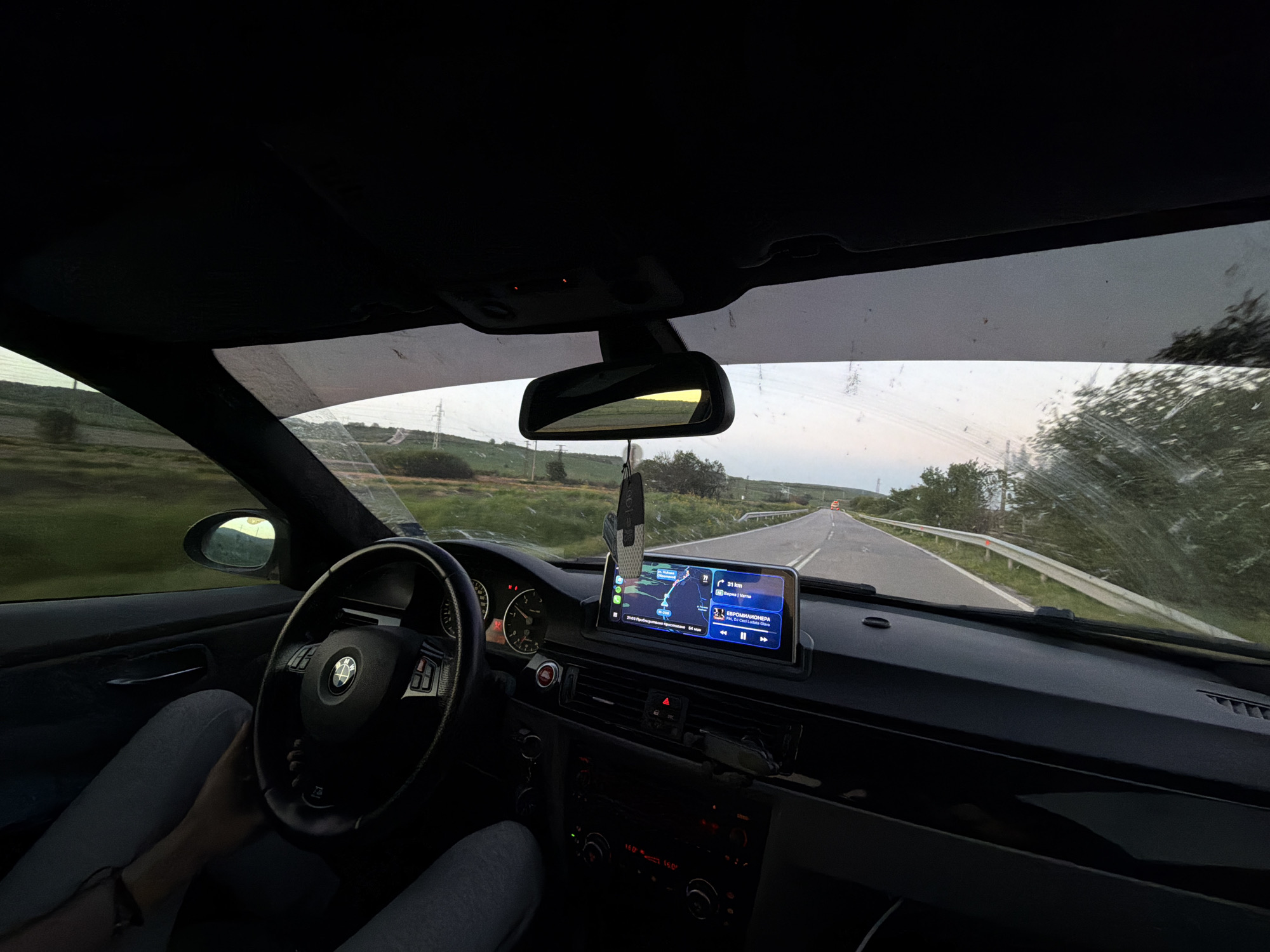 The image shows the interior of a car, specifically the dashboard, steering wheel, and part of the driver. The car is moving on a rural road, with green fields and some trees visible through the windshield. The GPS screen shows the car is 31 km away from Varna, Bulgaria. Based on this information, and the visible countryside, it is plausible that the car is in the vicinity of Boryana, Bulgaria, or on a road leading to or from it. The driver is holding the steering wheel, and wearing gray trousers. The car's dashboard has various controls and a navigation screen, and a decorative item hanging from the rearview mirror. The view from the windshield is slightly obscured by water marks. The light suggests it is either early morning or late evening.