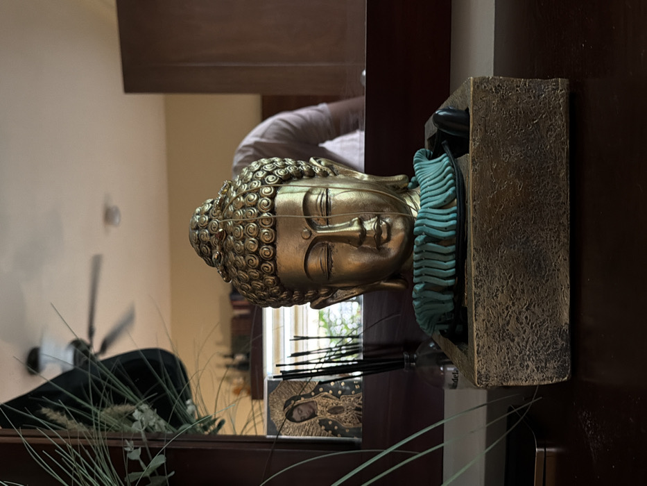 A gold Buddha head sculpture rests on a dark brown shelf. Turquoise elements accent the sculpture. Behind the sculpture, a mirror reflects the scene, showing a person in a white shirt and a ceiling fan. Plants and a religious icon are also visible in the reflection. A stone-like holder anchors the sculpture.