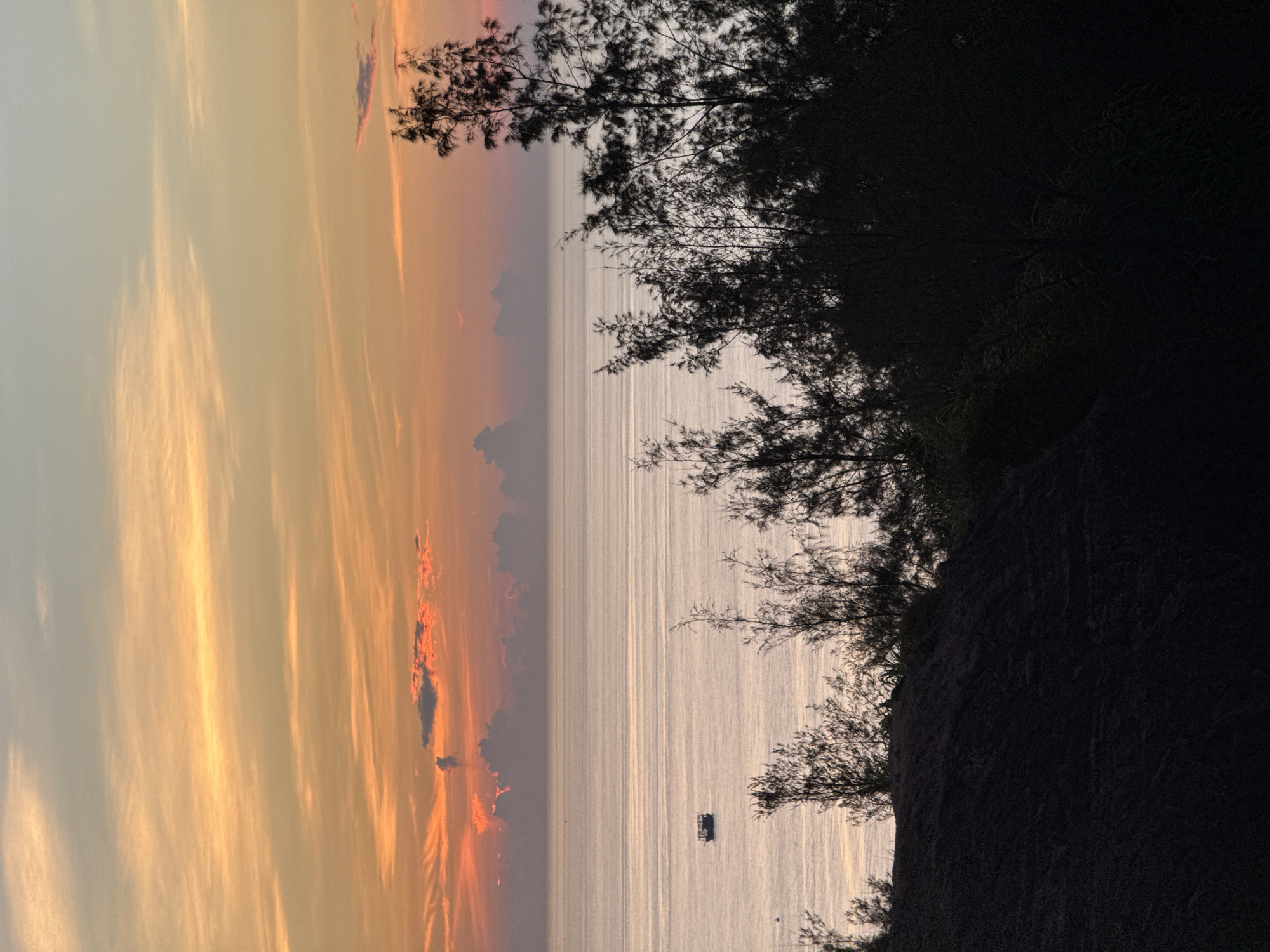 The image captures a serene seascape at dusk, with a warm, gradient sky of orange and pink hues stretching across the horizon. Thin, wispy clouds are scattered throughout the sky, reflecting the sunset's glow. In the distance, a silhouette of a small boat is visible on the calm, shimmering water. The foreground is dominated by the dark, silhouetted forms of trees and a sand dune, creating a stark contrast against the brighter sky and sea. The overall atmosphere is peaceful and tranquil, characteristic of a coastal sunset. The location is Lý Sơn Special Administrative Region, Vietnam.