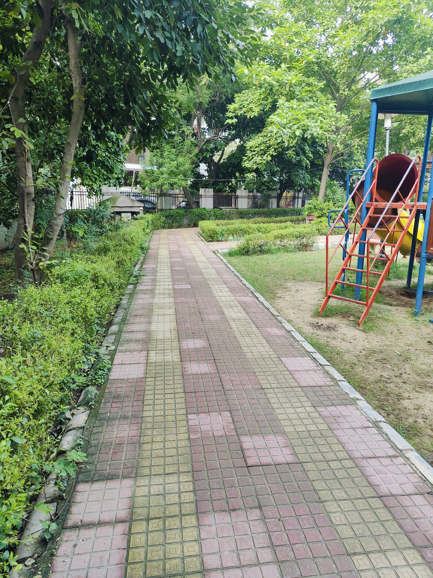 The image shows a paved walkway in a park setting in Gurgaon, India. The walkway is made of square tiles in alternating shades of pink and yellow, with a grey border. To the left of the walkway is a dense hedge of green bushes, and beyond that are trees and a fence. To the right of the walkway is a grassy area with a playground structure, including a blue and green slide with a red ladder. The playground equipment appears to be made of metal and plastic. The trees in the background are lush and green, suggesting a warm climate. The sky is overcast. There are no people visible in the image. The overall impression is of a well-maintained public space.