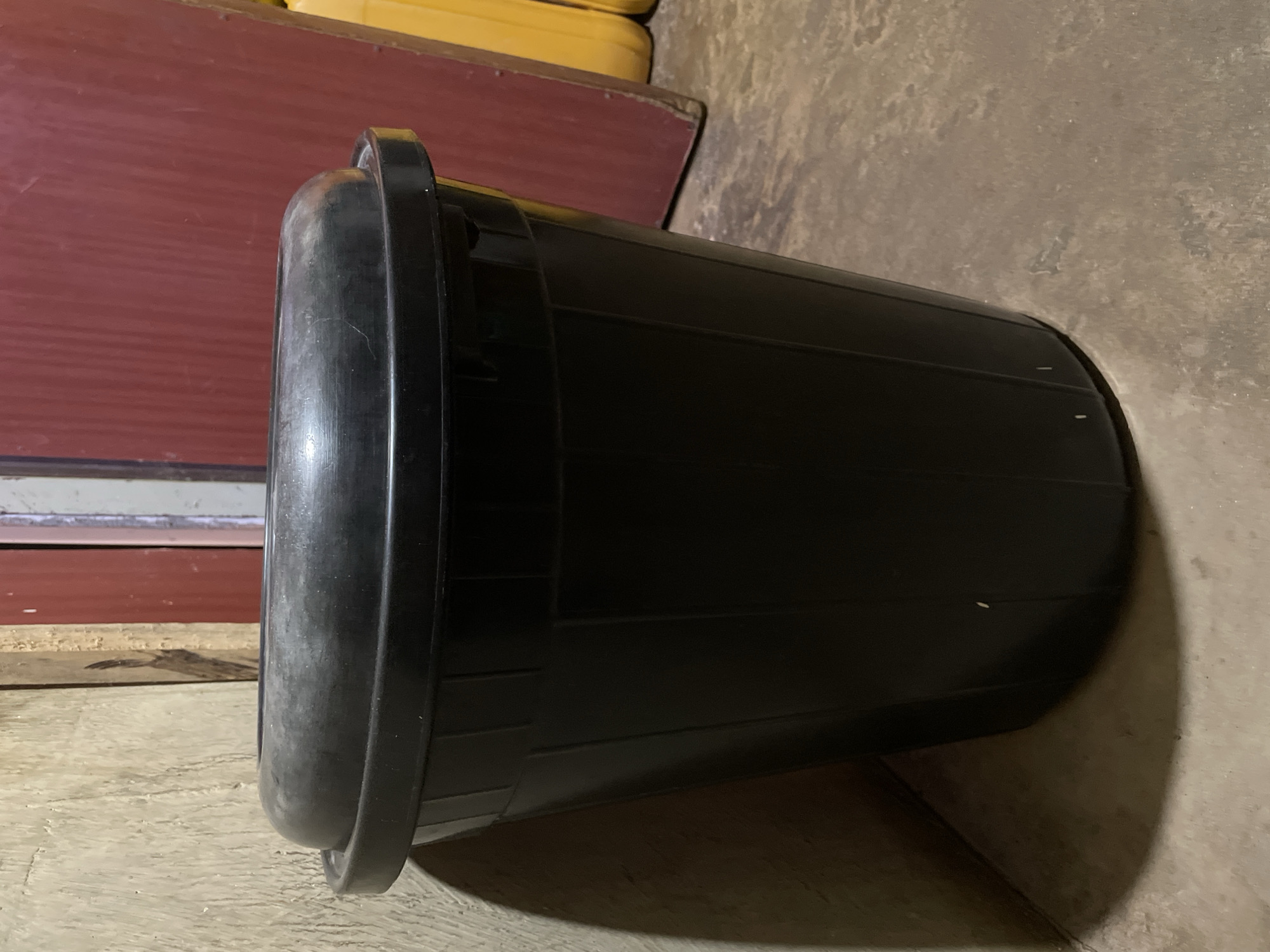 In Narayi, Nigeria, the image depicts a static indoor scene focused on a black plastic bin. The bin, positioned centrally, is cylindrical with a tapered body and vertical structural ribs. A matching black plastic lid, featuring a raised, domed center, is securely seated on the bin. Both the bin and its lid exhibit minor surface wear consistent with use.
To the immediate left of the bin, a textured, light-colored wall, possibly concrete or plaster, displays visible imperfections and a distinct curvilinear shadow cast by the bin. Further left and behind the bin, a vertical wooden element, likely a doorframe or support beam, is partially visible.
Behind the right side of the bin, a large, dark red vertical surface, consistent with a door or wall panel, shows signs of wear and possibly vertical grooves. In the far right background, partially obscured by the red surface, several yellow, rounded containers, possibly jerrycans, are visible, suggesting storage.
The bin rests on an uneven, multi-toned floor, appearing to be concrete or compacted earth, exhibiting varied texture and subtle debris. Ambient lighting casts shadows to the left and foreground of the bin, indicating a non-direct or somewhat dim light source. No individuals are present, and no actions or interactions are depicted. The overall environment appears utilitarian.