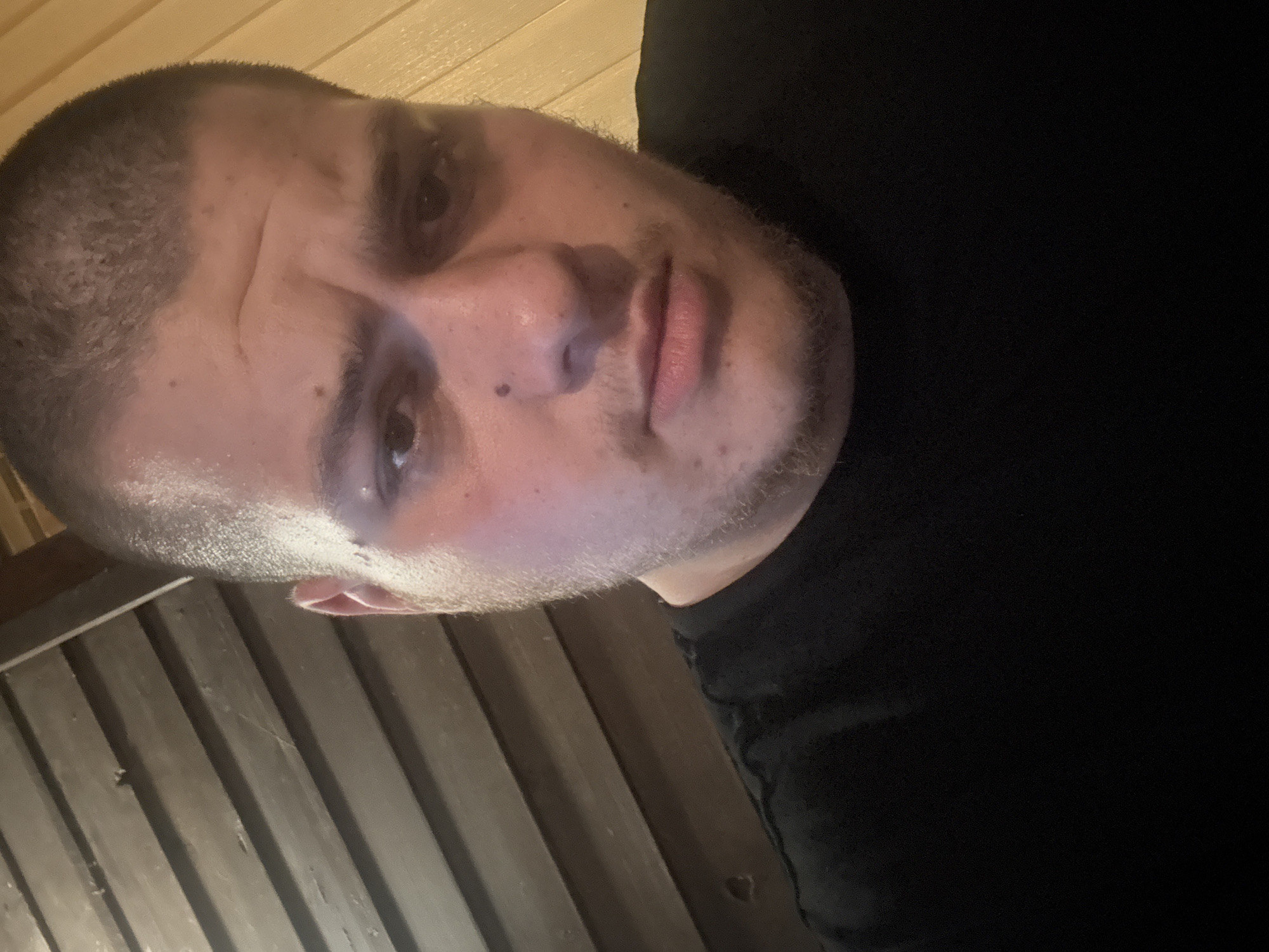 A male individual is depicted in a close-up, head-and-shoulders portrait shot, ostensibly taken in the city of Varshilo, Bulgaria. The subject is an adult male with a very short dark buzz cut and dark stubble on his face, including the chin and upper lip. He wears a plain black crew-neck t-shirt. His gaze is directed straight at the camera, with a neutral to slightly solemn expression, characterized by subtly furrowed brows and faint horizontal lines on his forehead. A small, dark, raised mole is visible on his left cheekbone (viewer's left). The background features two distinct sections of wooden paneling: dark brown horizontal planks on the left side of the frame and lighter, yellowish-tan vertical planks on the right. Interior lighting illuminates the scene, creating a slight sheen on the subject's forehead and right facial features.