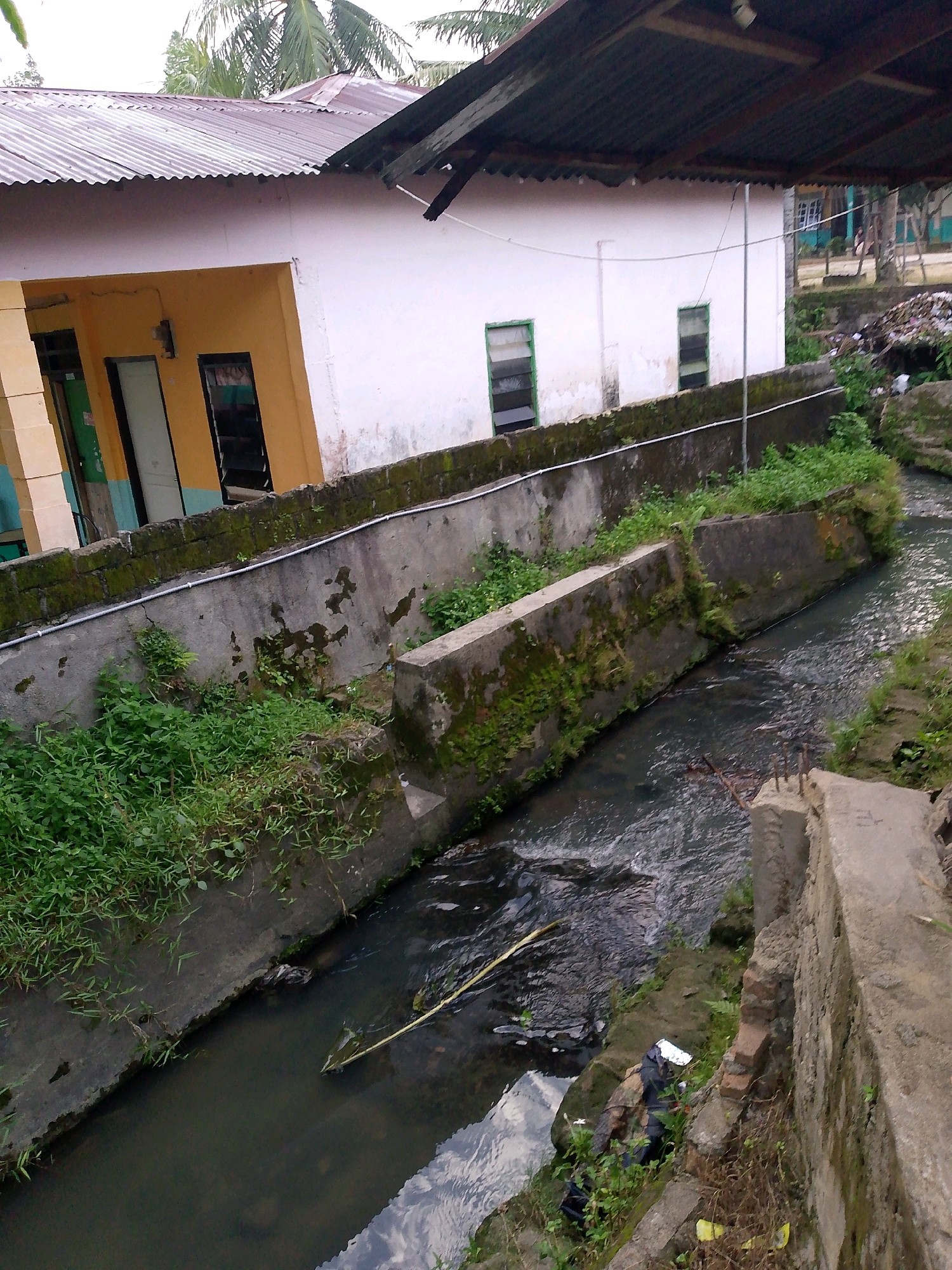 The image shows a polluted drainage canal running alongside a building. The building, which appears to be a school, has yellow and white walls and a corrugated metal roof. The canal is filled with murky water, debris, and overgrown vegetation. The surrounding environment appears to be rural or semi-urban, with palm trees visible in the background. The scene suggests a lack of proper waste management and environmental degradation. The location is identified as Dolok Merawan, Indonesia.