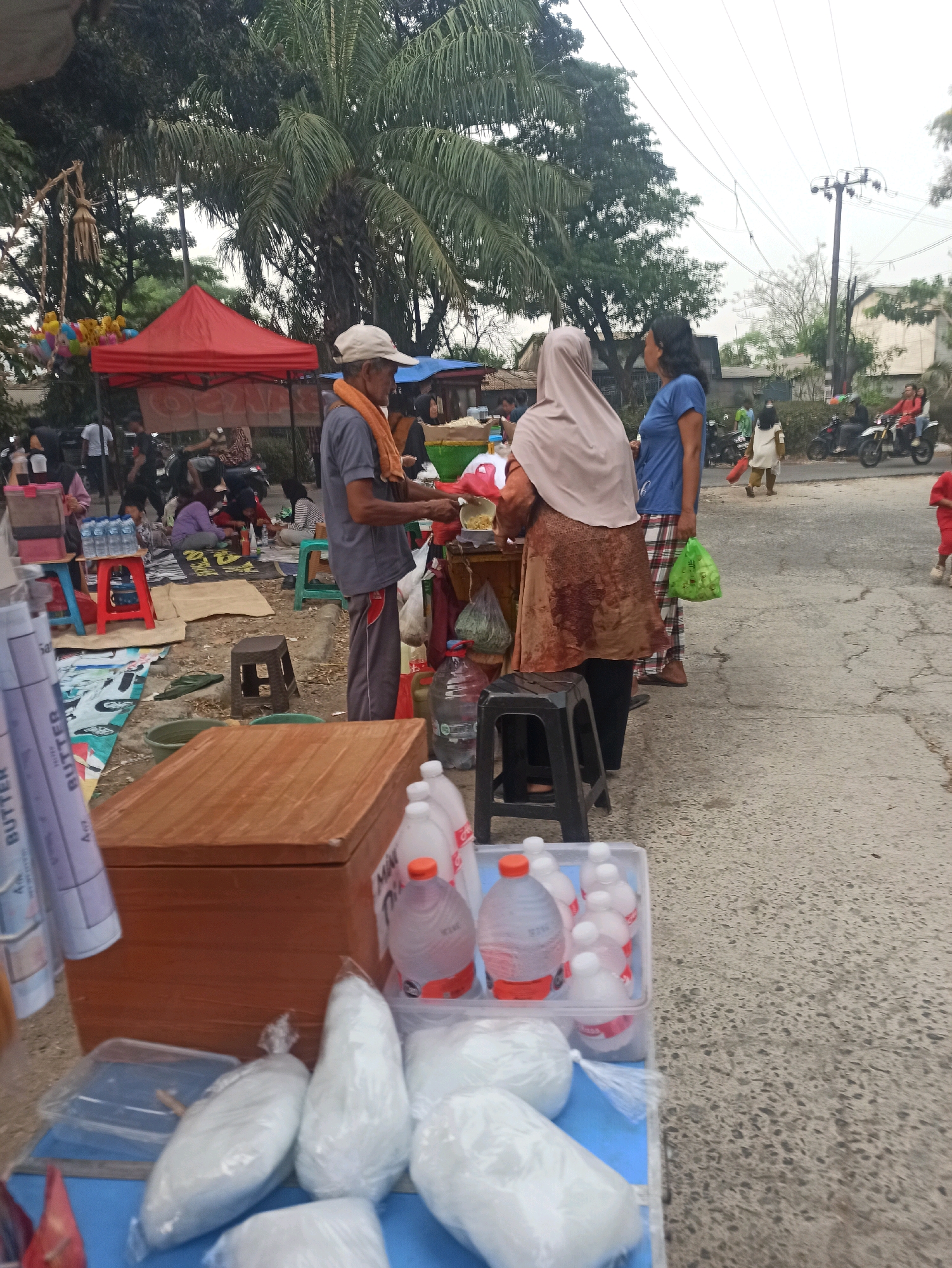 In Telagamurni, Indonesia, an outdoor market scene unfolds with a diverse array of goods and people. A vendor, wearing a light-colored hat and an orange towel around his neck, stands behind a stall, actively serving a customer who is wearing a beige hijab and a patterned dress. To their right, another woman in a blue shirt observes the interaction.
The market is set up on an asphalted ground, possibly a roadside, with various items displayed on the ground and on makeshift tables. In the foreground, a wooden chest serves as a counter for some of the goods, which include multiple plastic-wrapped bags of white powdery substances and several bottles of clear liquid with red and white labels. Nearby, a blue mat is spread out, holding more items like red and blue stools, and stacks of water bottles on a red plastic container.
Further back, a red tent provides shelter for more vendors and customers. Colorful balloons are visible hanging from a structure near the tent. In the background, a few motorcycles are parked, and some people are seated on mats, suggesting a lively marketplace atmosphere. A utility pole with wires stands tall against the overcast sky, typical of urban Indonesian scenery. The overall impression is one of a bustling local market, likely catering to daily needs.