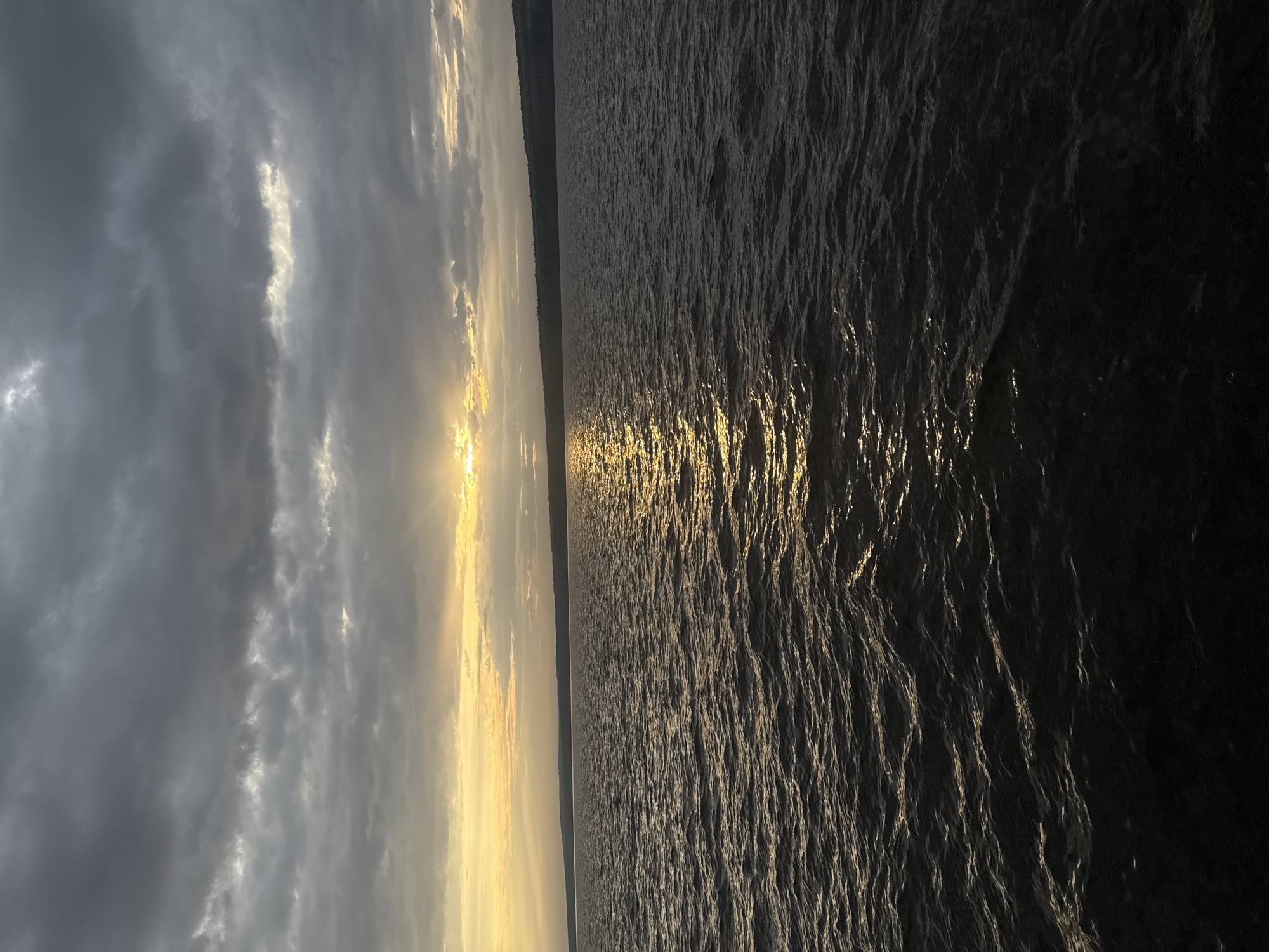 The image captures a sunset over the Kama River in Chaikovsky, Russia. The sky on the left shows a combination of dark grey clouds and bright streaks of sunlight. The sun, near the horizon, casts a golden glow across the scene. The water of the Kama River occupies the right half of the image. The surface is rippled, reflecting the sunlight as a shimmering golden path that stretches into the distance. In the middle distance, a dark line of vegetation or a distant shoreline is visible.