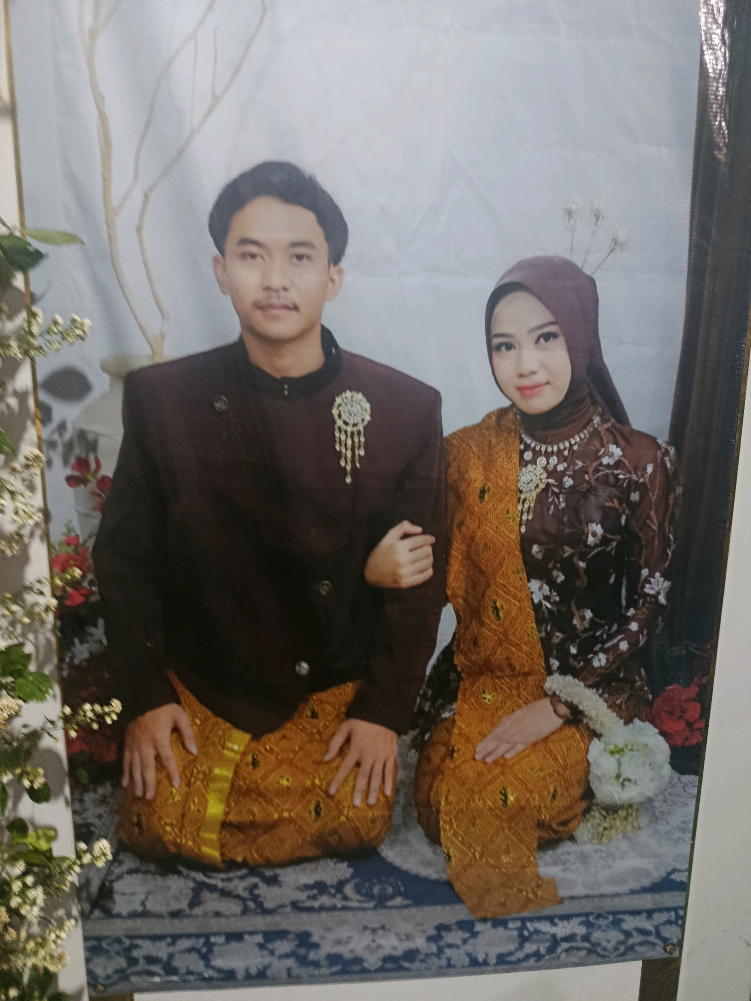 A studio portrait shows a young Indonesian couple posing for a photograph. The man, on the left, wears a dark brown, traditional jacket with intricate gold embroidery on the collar and a gold sash. A decorative brooch with pearls and dangling ornaments is pinned to his left lapel. He has short, dark hair and a neatly trimmed mustache. The woman, on the right, is dressed in a dark brown, patterned garment with floral designs. She wears a maroon hijab that covers her hair and neck. Her attire is accessorized with a pearl necklace and a large, ornate brooch similar to the man's. A golden sash, mirroring the man's, is draped across her chest and right shoulder. Both individuals are seated on a blue and white patterned rug. The backdrop features a pale, textured material, with a stylized light-colored tree branch on the left and a dark curtain on the right. Artificial flowers, including white blossoms, are visible to the left of the couple and a small bouquet is placed near the woman's hand. The lighting is soft and even, suggesting a controlled studio environment. The image is a framed photograph, likely taken in Karangkebagusan, Indonesia, given the traditional attire.