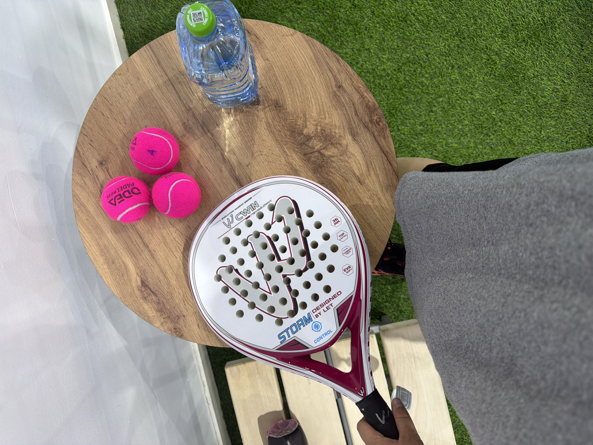A person is holding a padel racket with a white and pink design. The racket has "STORM CONTROL DESIGNED BY LET" printed on it. Three bright pink padel balls are resting on a round wooden table. Next to the balls is a clear plastic bottle of water with a green cap. The table is situated on a patch of artificial grass, with a white wall in the background. The scene appears to be outdoors, possibly in Chaikovsky, Russia, given the context provided. The person's legs and feet are visible in the foreground, wearing grey sweatpants and black athletic shoes. The lighting suggests it is daytime.