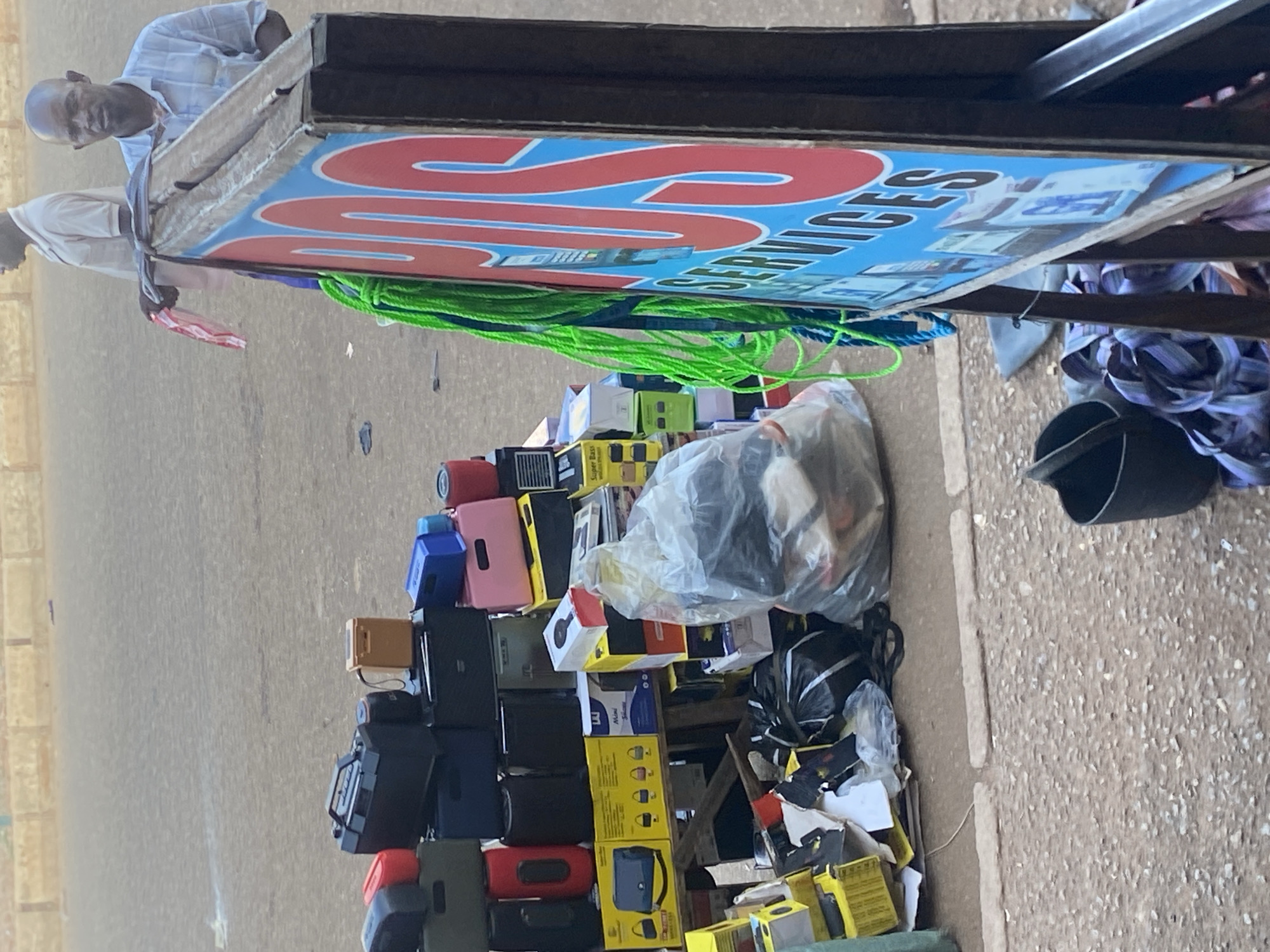 An outdoor street stall in Bukuru, Nigeria, displays various electronic goods, including portable speakers and radios, stacked on wooden shelves. In the foreground, a sign with "VDS SERVICES" written in red and blue lettering is prominently displayed. Two men are visible in the background; one is standing behind the stall, and the other is further back, seemingly a customer. The ground is paved with asphalt and gravel. A collection of plastic bags, some filled with items and others empty, are scattered around the stall. Green ropes hang from the side of the stall.